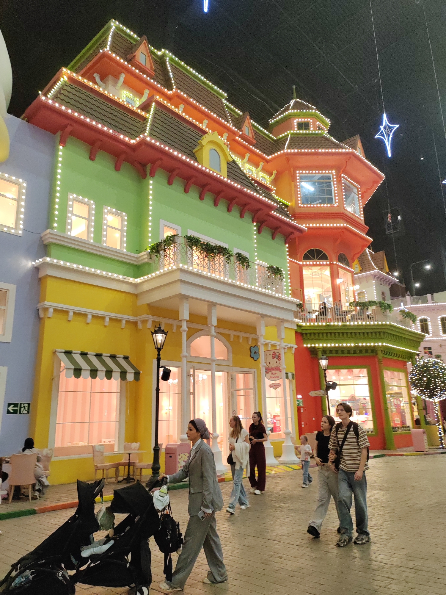 The image captures an outdoor scene at what appears to be an amusement park or entertainment venue, likely indoors given the dark ceiling and artificial lighting. The architecture is whimsical and brightly colored, resembling cartoonish buildings adorned with numerous strings of white lights. The dominant colors of the buildings are pastel blue, vibrant green, bright yellow, and vivid orange, with red trim. Balconies with railings and decorative plants are visible on the upper levels.
In the foreground, a woman in a grey pantsuit and headscarf is pushing a double stroller. The stroller is black and appears to contain one or two young children. To the right of the woman, a group of four individuals, two young women and two young men, are walking together. One of the young women has her arm around the other. Further in the background, other people can be seen seated at tables and walking.
A street lamp with a black pole and ornate lantern stands near the woman with the stroller. In the upper right portion of the image, a blue star-shaped light fixture hangs from the ceiling. The ground is paved with interlocking bricks. The overall impression is a festive and cheerful atmosphere.