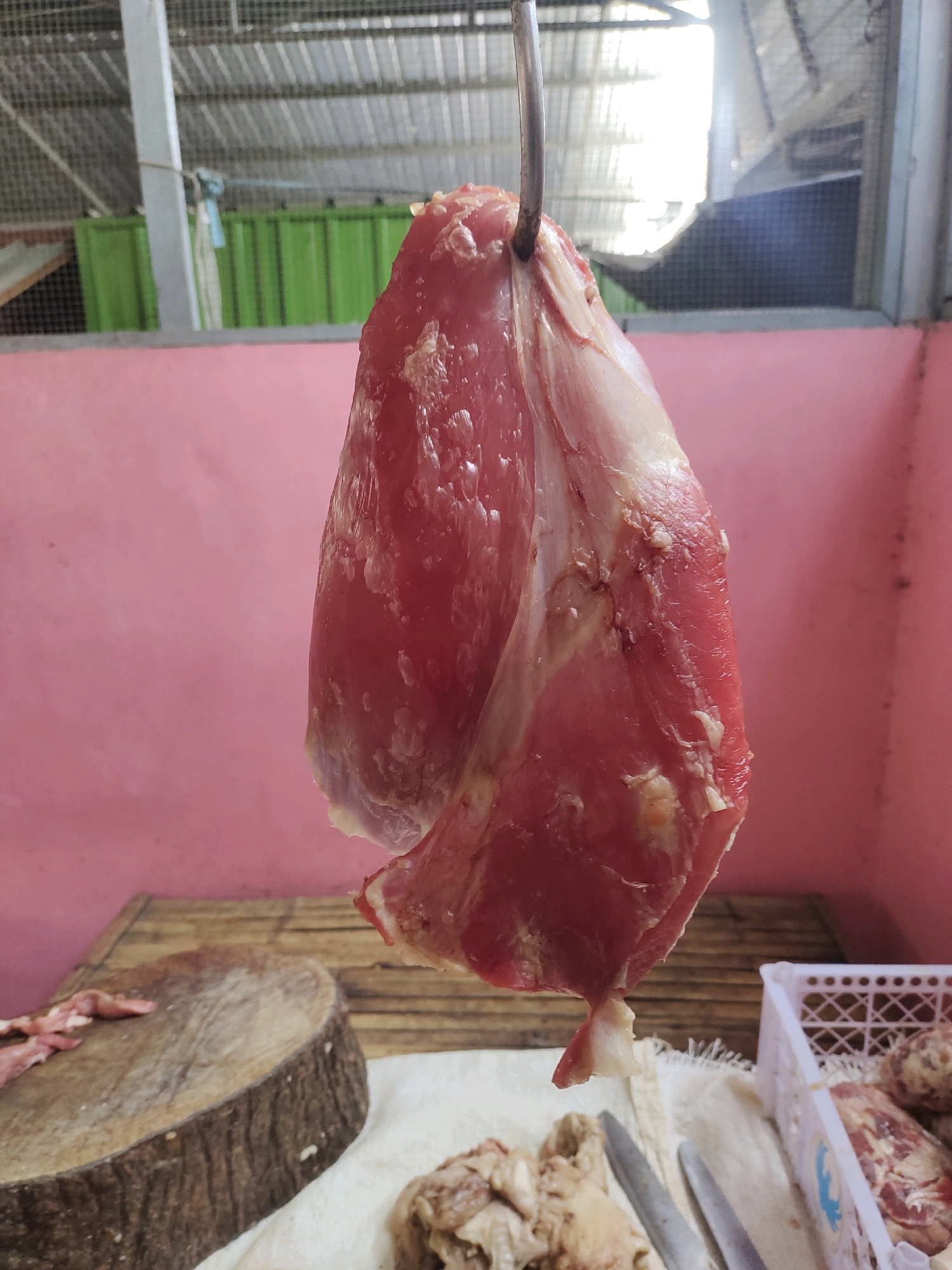 A side view medium shot captures a large cut of raw, red meat suspended by a metal hook from an overhead bar. The meat is plump and appears to be beef, with visible streaks of fat and connective tissue. The background consists of a pink wall, a wooden surface, and a white cloth. To the left of the meat, a round wooden chopping block is visible, along with a few smaller pieces of raw meat. To the right, a white plastic basket holds more meat. The scene is likely set in a market or butcher shop in Sambi, Indonesia, given the open-air construction and typical market elements. The lighting suggests it's daytime, with bright light coming from the upper right.