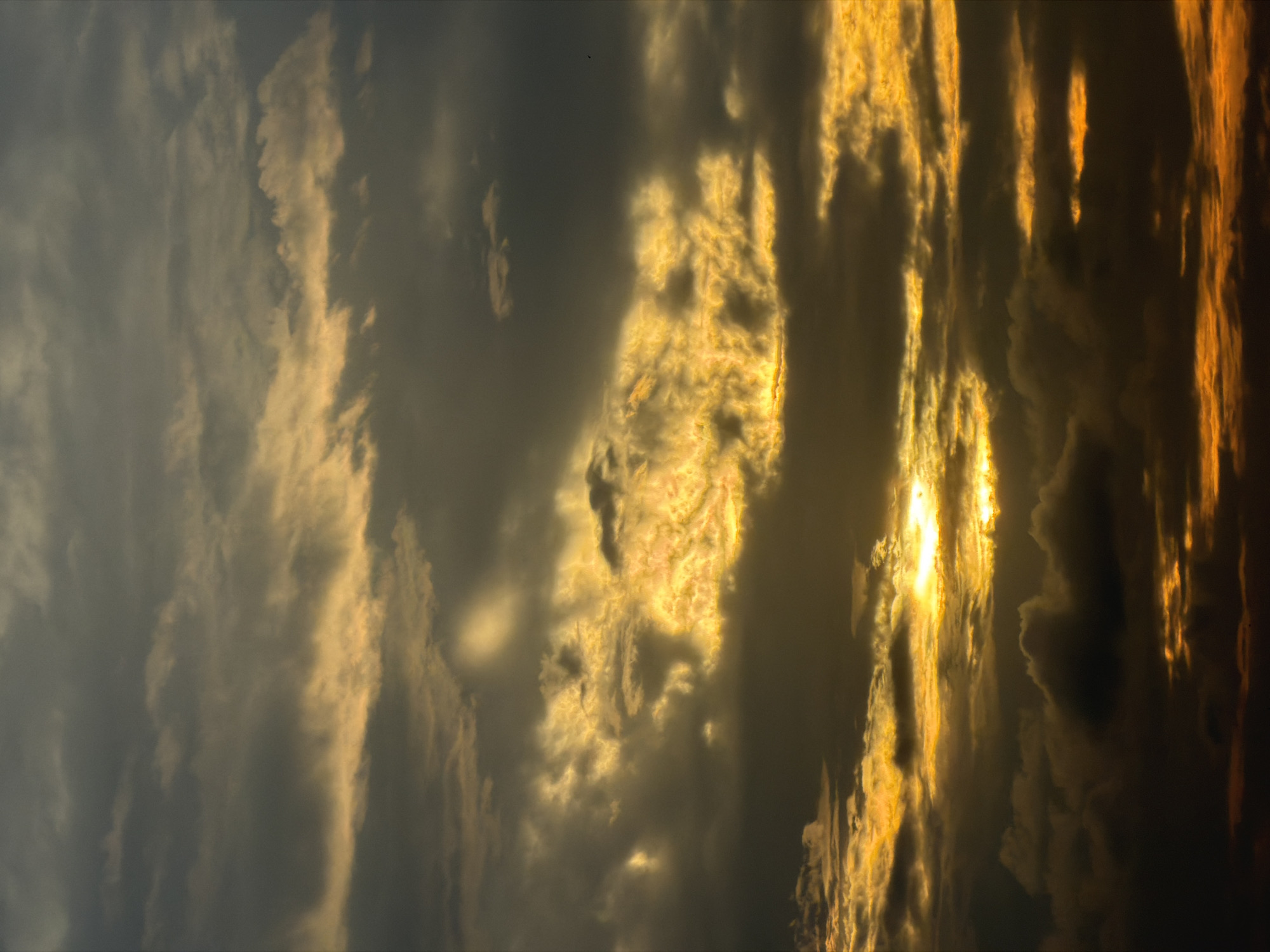 This is a photograph of a dramatic sunset over the clouds in North Kuta, Indonesia. The sky is filled with layers of clouds, some dark and ominous, others illuminated by the setting sun with hues of yellow, orange, and gold. The sunlight pierces through the clouds, creating an ethereal glow and highlighting the textures of the cloud formations. There are no visible people, subjects, or objects in the frame, focusing solely on the atmospheric phenomenon. The environment is entirely the sky, captured from a perspective that emphasizes the vastness and dynamic nature of the clouds at dusk.