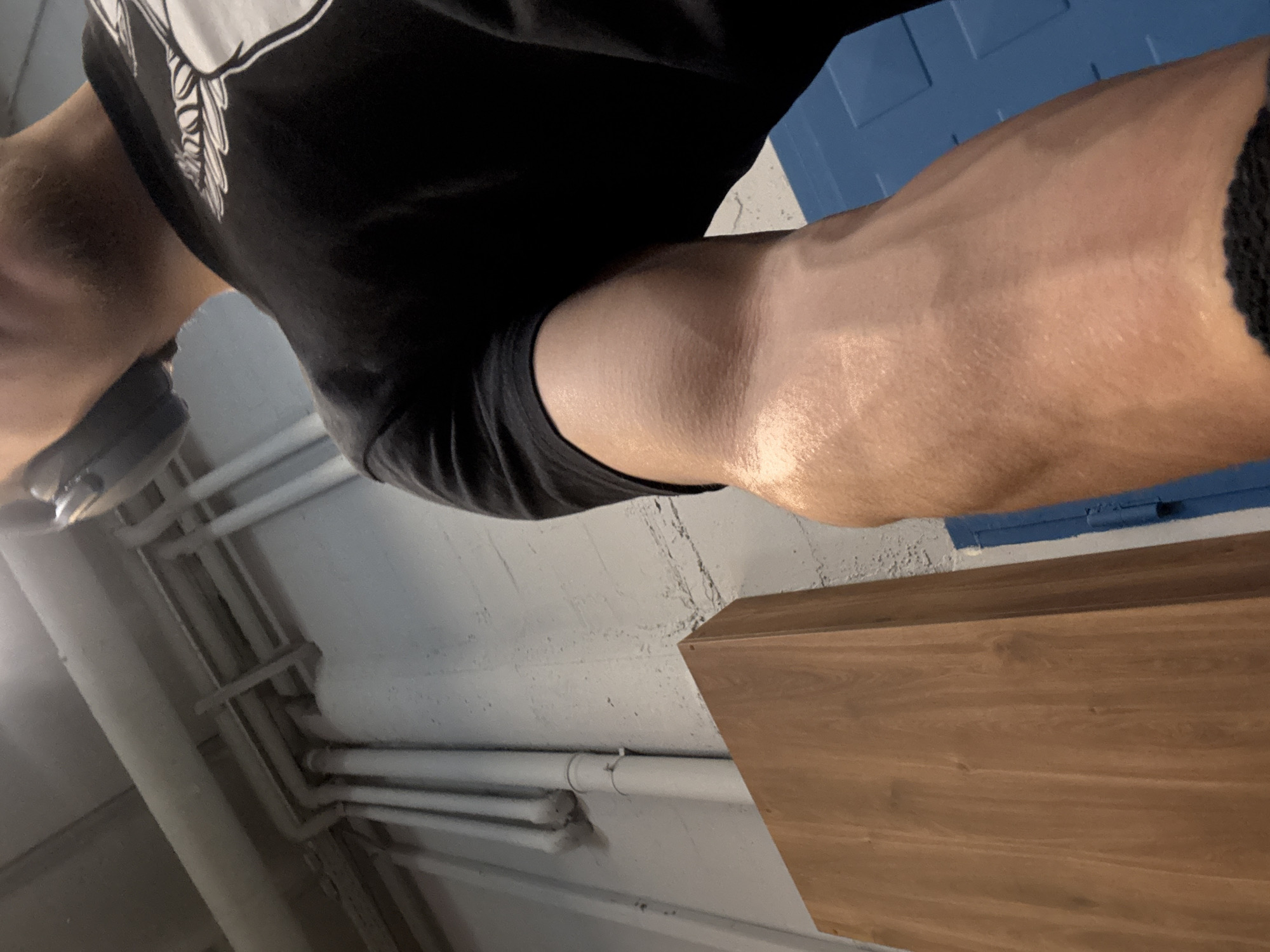 The image shows a person's leg, bent at the knee, with a black shirt and shorts. The person is wearing something on their head, possibly headphones or glasses. The background consists of a wall with pipes running along it, a wooden surface, and a blue painted area. The location is not identifiable as Sofia, Bulgaria from the image alone.