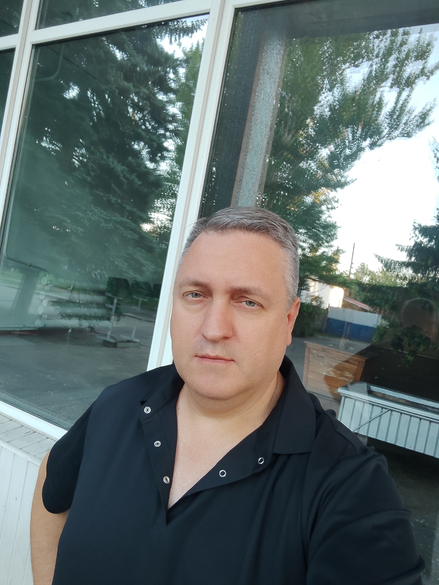 A man with graying hair and a fair complexion is taking a selfie in front of a large window. He is wearing a black collared shirt with silver snaps. The window reflects trees and buildings. The man is facing the camera and looking directly at it with a neutral expression. The overall lighting suggests it is daytime, possibly late afternoon or early evening.