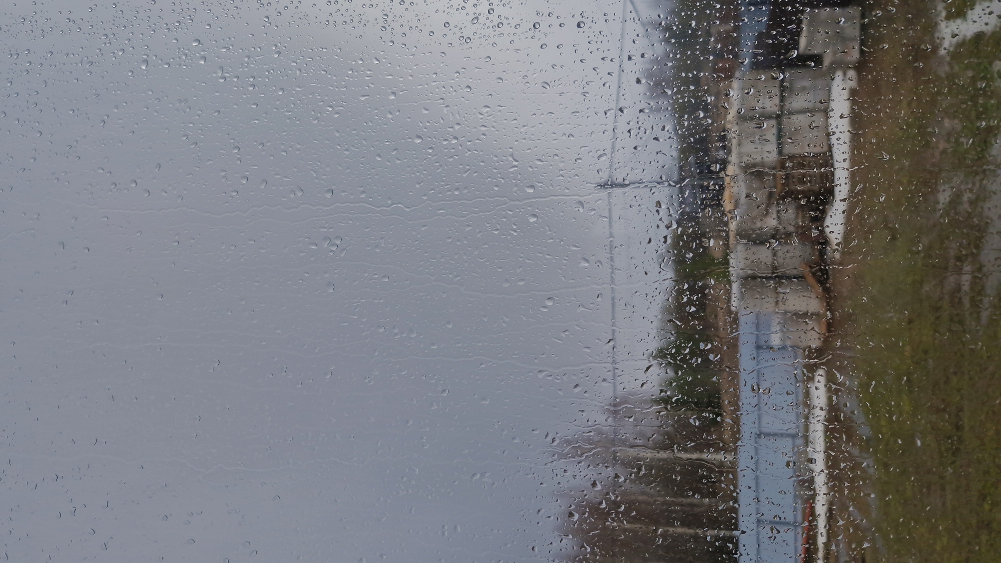 The image shows a view from behind a rain-streaked window. Outside the window, on the right side of the frame, there is an out-of-focus view of a street or sidewalk, with possible buildings and trees. The location is likely Otchor, Russia. The buildings appear to be of a generic, possibly Soviet-era design. The sky is overcast, indicating rainy weather. There are no people visible in the image. The overall impression is one of a bleak and wet day.