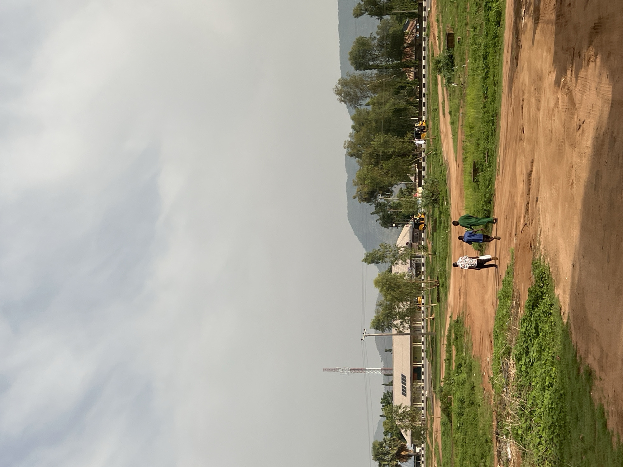 The image depicts a scene in Howayi, Nigeria, featuring a dirt road running parallel to a green strip. Three people are walking on the road. The background includes buildings, trees, a radio tower, and a mountain range. The sky is overcast.