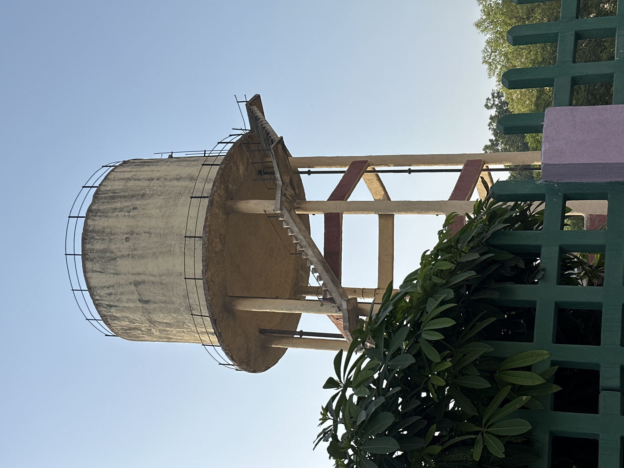 The image features a concrete water tower against a clear blue sky. The tower is cylindrical with a ladder leading to the top. It is supported by concrete beams, some reinforced with red paint. To the right of the tower, a green fence with square cutouts is partially obscured by leafy plants. The location is likely Mathura, India. There are no people visible.