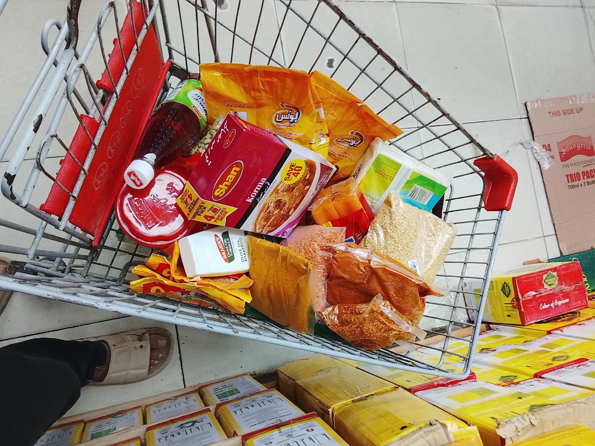 A shopping cart filled with various grocery items sits in what appears to be a store or market. The items include packets of "Bonus" snacks, "Shan Korma" seasoning mix, "Palmer's Olive Oil Formula" shampoo, lentils, spices, and other unidentified products. Boxes with yellow packaging are stacked underneath the cart.
A person's foot, clad in a black sock and a sandal, is visible near the cart.
A cardboard box labeled "Shangrila The Food Experts TRIO PACK" is visible in the background. A red box with the label "Colour of Happiness" is also present.
The OCR suggests the products are for sale in Pakistan, indicating the location is likely within that country. Given the information available, it is not possible to confirm the exact location in Gulgasht Colony.