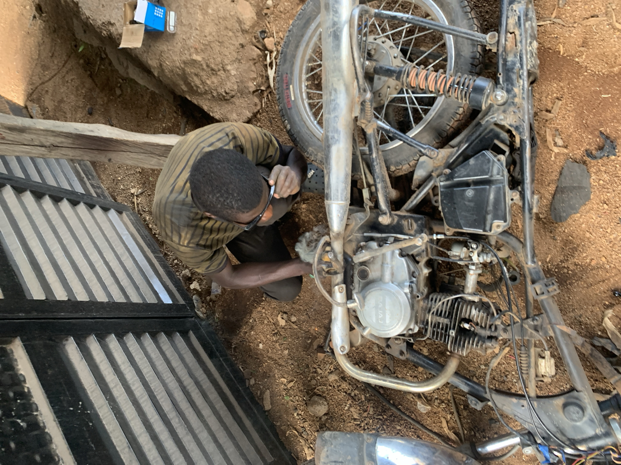 A man is working on a motorcycle on a dirt ground. He is wearing a striped shirt and dark pants, and glasses. The motorcycle is disassembled, with parts spread around. The scene appears to be outdoors, possibly in a rural area, with a large rock and wooden structure visible in the background. The ground is covered in dirt and small stones, and there are some pieces of debris. There is no clear indication of the time of day or weather. The man's focus seems to be on the engine of the motorcycle. There is a blue and white box with some items on top of it behind the man. The words "BAJAJ" are visible on the engine. The image is taken from a high angle looking down at the man and the motorcycle.
