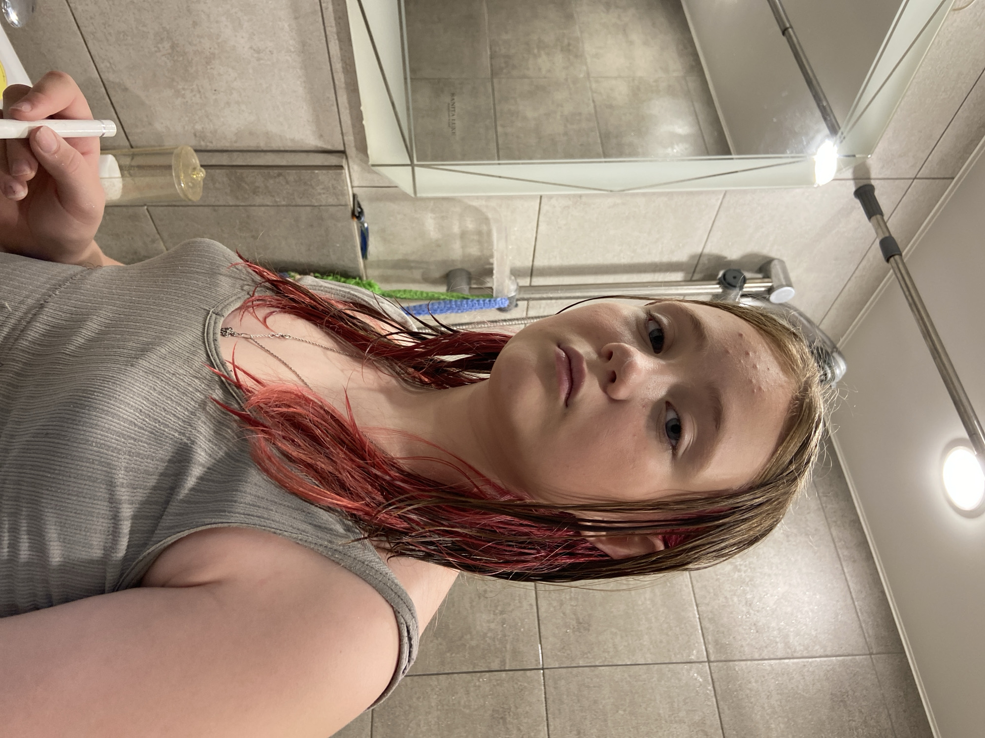 The image shows a young person with wet, reddish-brown hair in a bathroom. The person is wearing a grey ribbed tank top and a silver necklace. To the left, a mirror reflects the bathroom light and part of the room. A shower head and faucet are visible in the background, along with tiled walls. A razor and a white object, possibly a toothbrush or applicator, are on the counter in the foreground. The overall setting appears to be a domestic bathroom, consistent with a residential location in Chaikovsky, Russia.