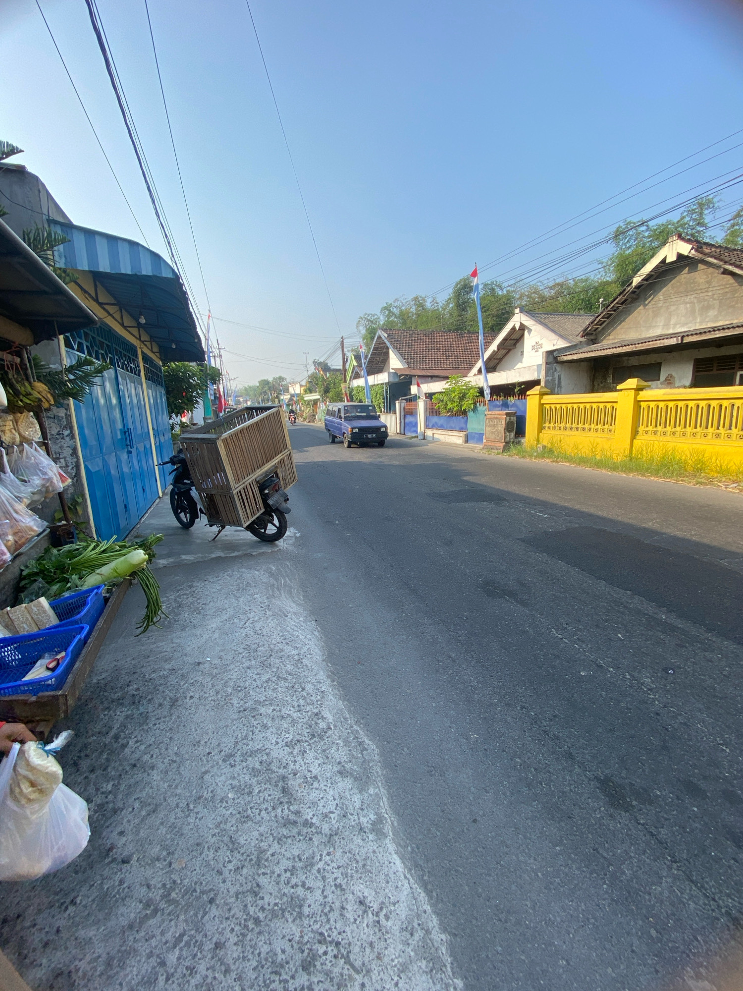The image depicts a street scene in Gurah, Indonesia, on a clear day with a bright blue sky. On the left side of the asphalt road, a small market stall displays produce, including leafy green vegetables and what appear to be bananas. A blue tarp covers the stall. Next to the stall, a motorcycle is parked, equipped with a large wooden cage on its rear. A person's hand is visible in the lower left corner, holding a white plastic bag. The road itself shows signs of wear. On the right side of the road, houses with tiled roofs are visible, separated by a bright yellow fence. A light blue pickup truck is driving away in the distance on the right side of the road. Indonesian flags on poles are scattered along the street. The overall impression is a typical street view of a residential and commercial area in Indonesia.