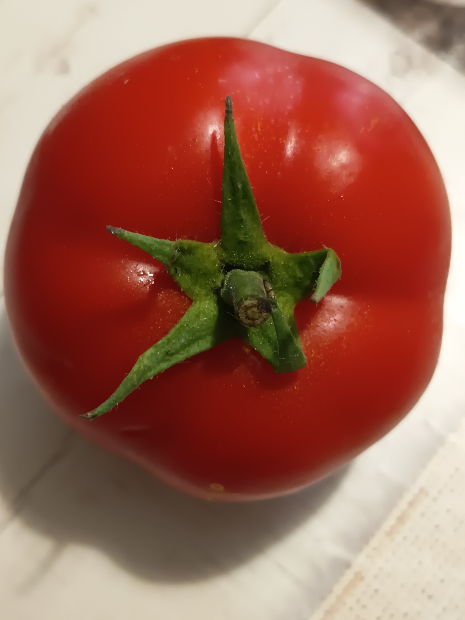 A close-up, overhead shot of a single, ripe red tomato is presented against a lightly patterned, off-white surface. The tomato occupies the majority of the frame, with its stem and green calyx clearly visible at the top center. The surface beneath the tomato appears to be a table or countertop, with subtle shadows indicating the direction of light. The image provides no information about the city of Izmail, Ukraine, or any people or actions.