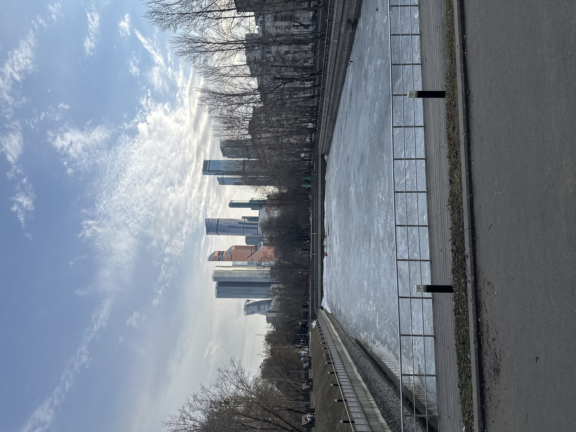 The image depicts a wide-angle view of a frozen body of water within a city park, with a modern skyscraper skyline in the background. This location is identified as Presnensky Pond, situated adjacent to Krasnopresnensky Park, with the Moscow International Business Center (Moscow City) visible in the background, in Moscow, Russia.
In the foreground, a dark asphalt road spans the bottom, bordered by a concrete curb. Beyond the curb is a narrow strip of brown soil and sparse vegetation, followed by a wooden boardwalk equipped with silver-colored metal railings and black bollard-style lights with white tops.
The midground is dominated by a rectangular body of water, completely covered by a sheet of ice, indicating freezing temperatures. A small red object and a small dark object are visible on the ice surface. The water's banks are lined with concrete retaining walls and sloped sections of dark stones. Along the left bank, another elevated concrete pathway and a grassy hill are visible, featuring several park benches, on which a few individuals are seated. Numerous bare deciduous trees with dark branches line both sides of the water channel, suggesting late autumn, winter, or early spring.
The background reveals the distinct skyline of the Moscow International Business Center (Moscow City), comprising multiple towering skyscrapers of varying contemporary architectural designs and reflective glass facades. Notable buildings include structures with blue, silver, and distinctive orange-tinted glass. The sky above is partly cloudy, displaying patches of blue sky amidst scattered white and grey clouds. No significant actions or interactions are clearly discernible among the few individuals present.