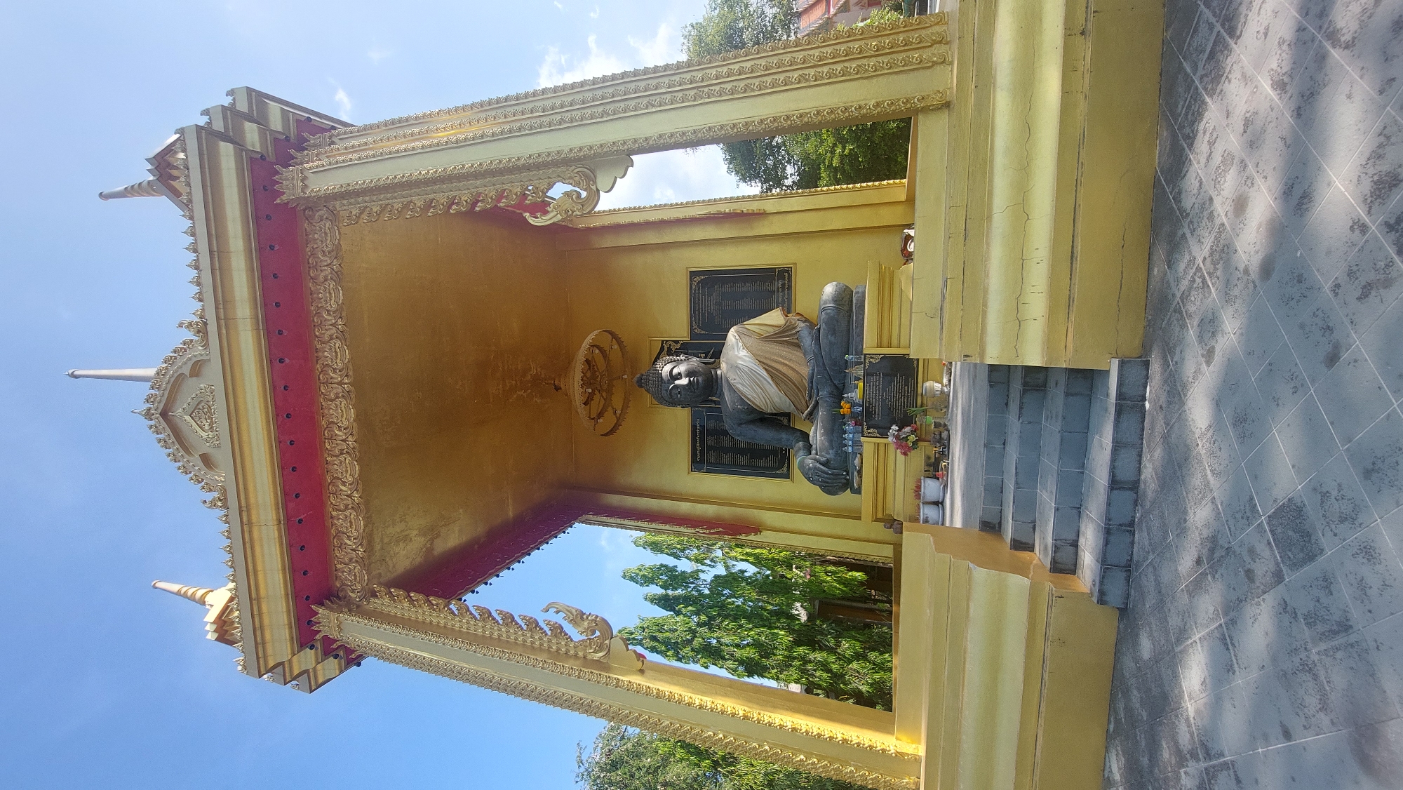 The image displays a serene outdoor shrine in Baan Mae Nam, Thailand, centered around a large, dark grey Buddha statue. The Buddha, draped with a light-colored sash, sits in a meditative pose, flanked by small offerings of flowers and what appear to be incense holders. This sacred figure is housed within an ornate pavilion, predominantly golden with vibrant red accents and intricate decorative carvings typical of Thai architectural style.
The pavilion features a multi-tiered, elaborate roof with pointed finials, set against a clear blue sky with sparse white clouds. Lush green trees are visible through the openings of the pavilion, highlighting its outdoor setting. Below the shrine, a series of grey steps leads up to the golden platform where the Buddha rests, and a tiled ground extends outwards, showing distinct shadows from the bright daylight. Two framed plaques with unreadable Thai script are mounted on the golden wall behind the Buddha statue. The scene suggests a tranquil place of worship or cultural significance, bathed in the strong light of day. No people are visible.