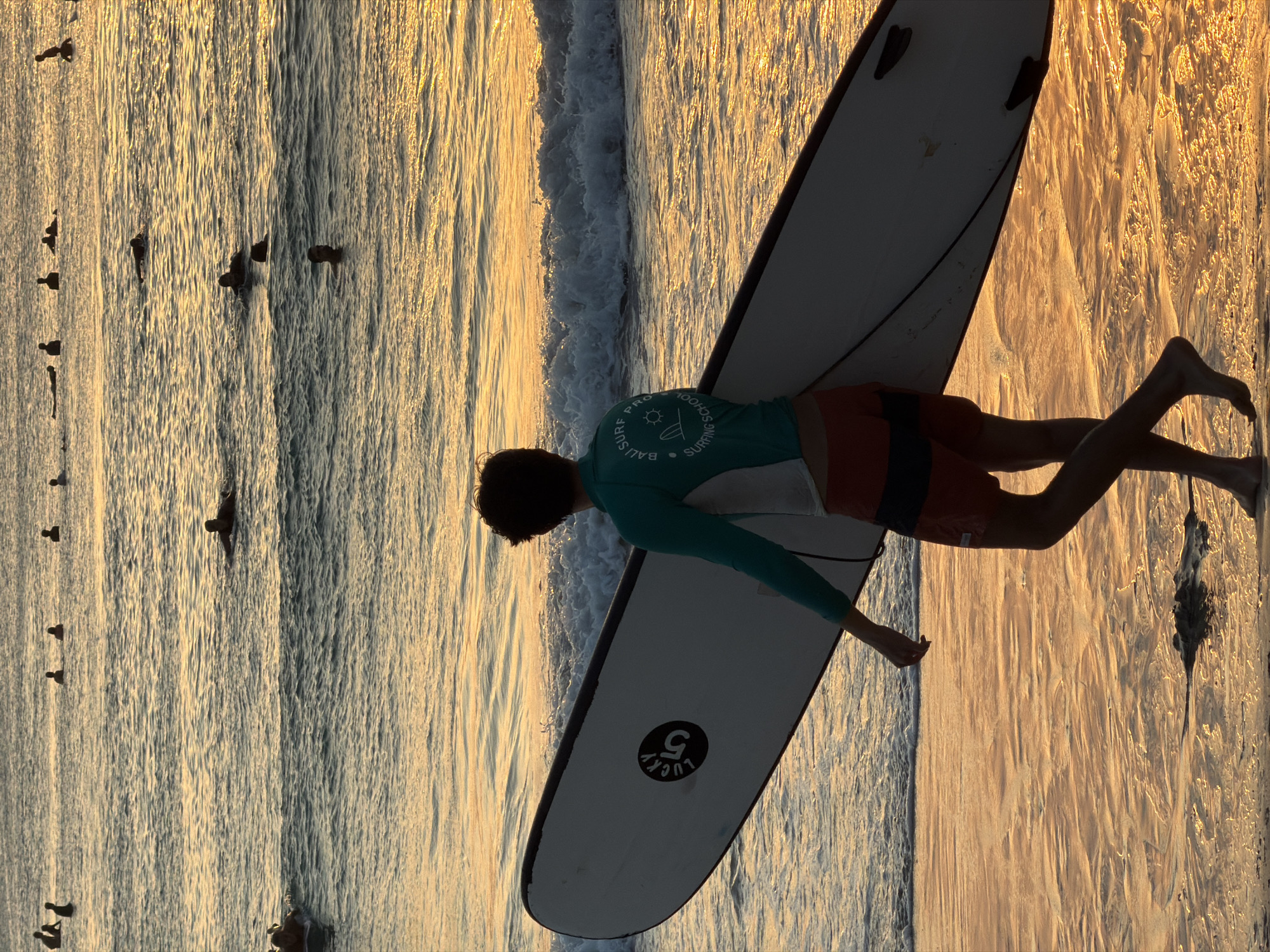 A young person walks along the beach in Kuta Selatan, Indonesia, carrying a white surfboard with a black stripe and a circular logo that reads "LUCKY 5". The person is wearing a teal long-sleeved rash guard with a white panel on the chest and orange and black striped shorts. They are walking towards the ocean, which is filled with many surfers paddling and sitting on their boards. The sun is low in the sky, casting a warm golden light on the water and the wet sand. The waves are small and gentle. In the distance, more surfers are visible, some further out in the ocean. The sky above is a soft gradient of colors, indicating either sunrise or sunset. The overall scene depicts a serene and active beach environment.