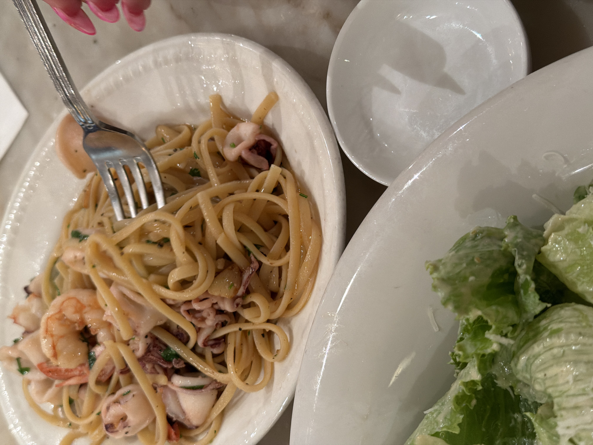 The image shows a meal, likely in Tel-Aviv, Israel given the user's request. The meal consists of a plate of seafood linguine with shrimp and squid, a bowl of Caesar salad, and a small empty bowl. A fork is piercing the linguine. A hand with pink nail polish is visible near the fork. The scene is likely indoors on a light-colored surface.