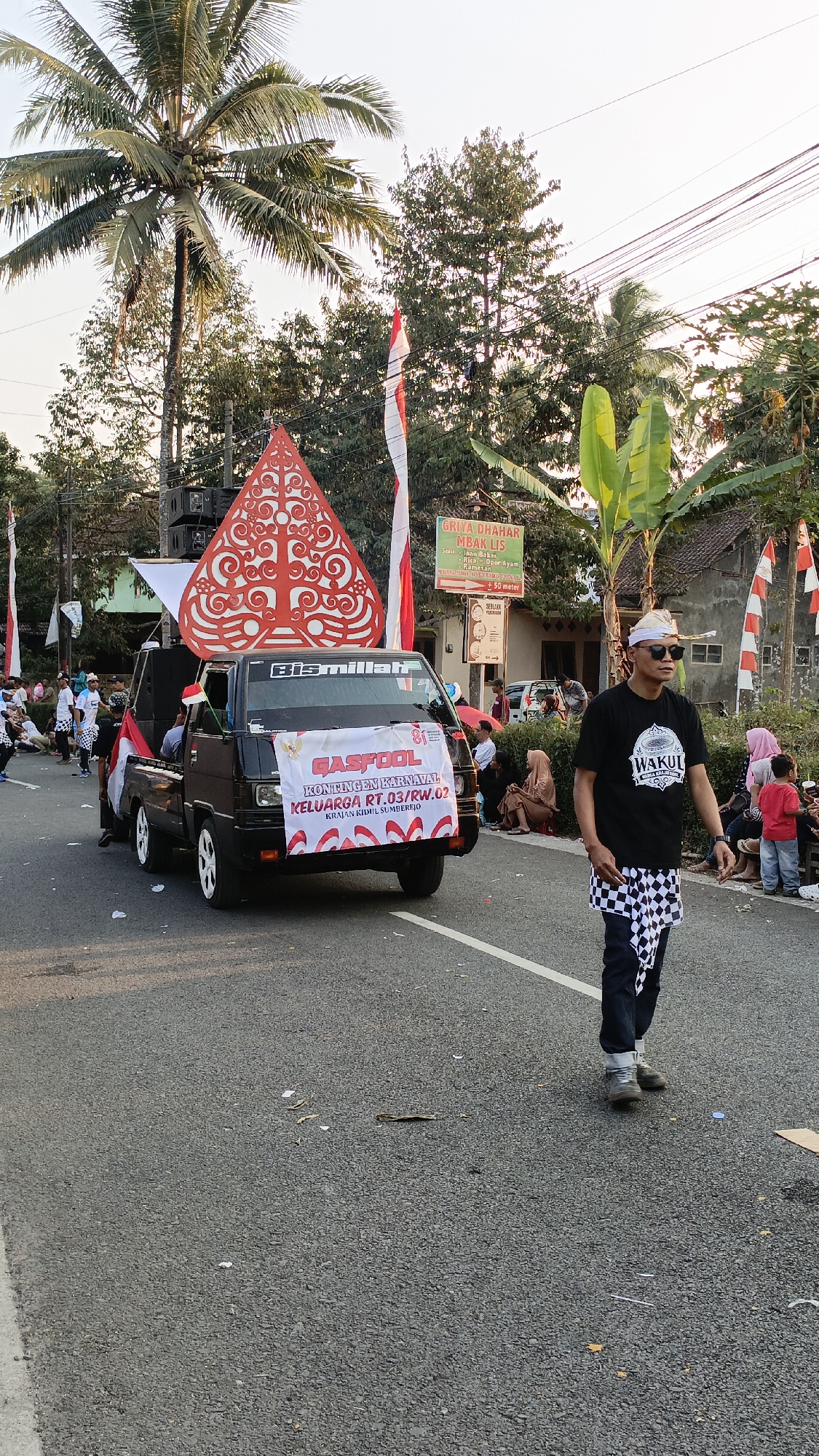 The image depicts a street scene in Krajan Lor, Indonesia, during what appears to be a parade or cultural event. In the foreground, a man is walking on the right side of the road, wearing a black t-shirt with a white logo, sunglasses, a head covering, and a checkered sarong or cloth around his waist. He is looking to his right, away from the camera.
To the left of the man, a black pickup truck is participating in the event. It is adorned with Indonesian flags and a large, red and white traditional Javanese puppet (wayang golek) figure on top. A banner on the front of the truck reads "GASFOOL KONGENEN KARNAVAL KELUARGA RT.03/RW.02 KRAJAN KIDUL SUMBERJO," suggesting it represents a neighborhood group from Krajan Kidul, Sumberjo. The truck also has a speaker system mounted on it.
Several people are visible on the sidewalks and along the road, spectating the event. Some are seated, while others are standing. The environment includes trees, power lines, and buildings in the background, indicating a semi-urban or rural setting. The lighting suggests it is daytime, likely late afternoon due to the warm, golden light. The road is paved and has some debris scattered on it.