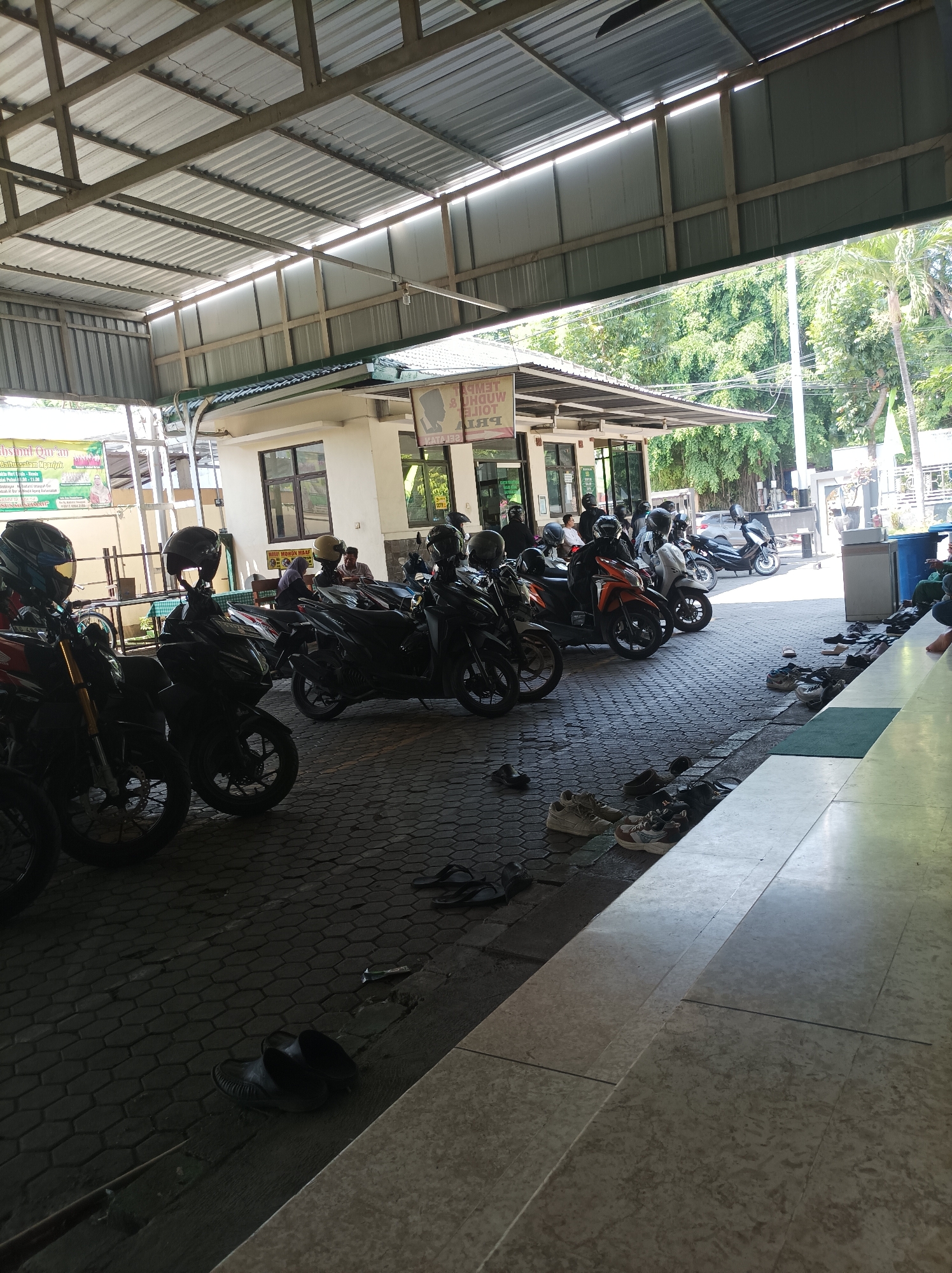 The image depicts an outdoor covered area, likely part of a public building or institution in Nganjuk, Indonesia. A row of motorcycles is parked under a metal roof supported by concrete pillars, suggesting a parking area for visitors or staff. Several people are seen on or near the motorcycles, some wearing helmets. The ground is paved with interlocking hexagonal bricks. To the right, there is a raised tiled floor area, possibly an entrance or walkway, with several pairs of footwear (sandals and shoes) neatly arranged along the edge. Beyond the covered area, lush green trees and foliage are visible, indicating a tropical or subtropical climate. The architecture appears functional and practical, with visible corrugated metal roofing and simple building structures. The overall scene suggests a place of gathering or daily activity, where people arrive by motorcycle and disembark before entering a building.