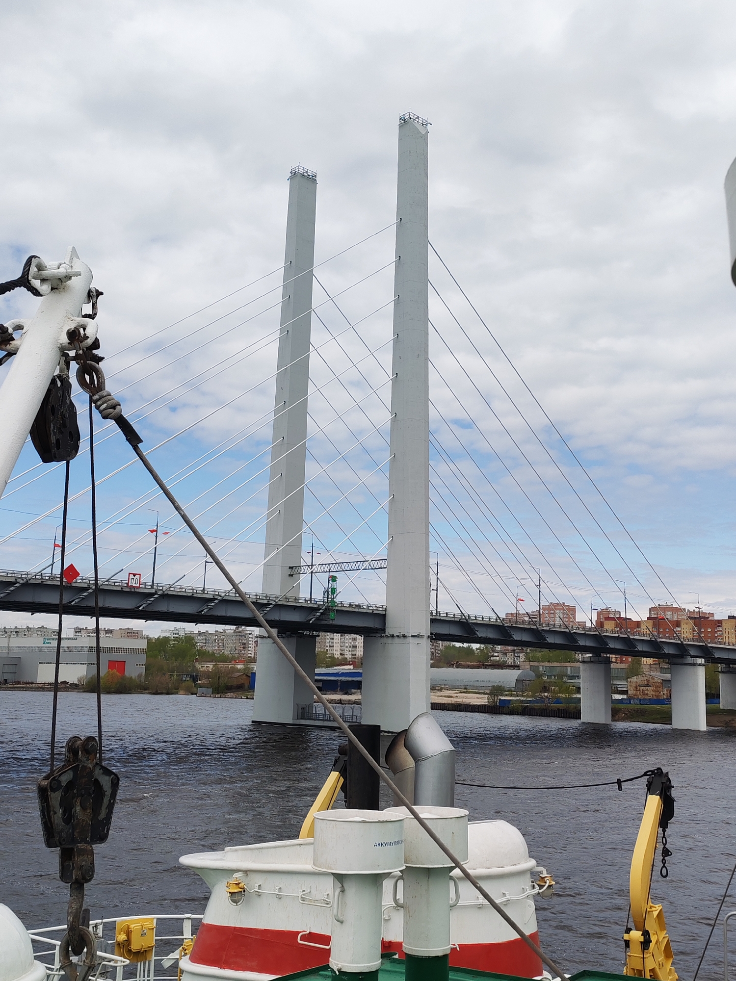 The image shows a bridge with two tall, gray towers supporting it with cables. The view is taken from what appears to be a boat or ship, as evidenced by the presence of maritime equipment in the foreground. The water surface is dark, and the background includes buildings and greenery on the shore. The sky is cloudy with patches of blue. The location appears to be urban, possibly near a waterway in a city.