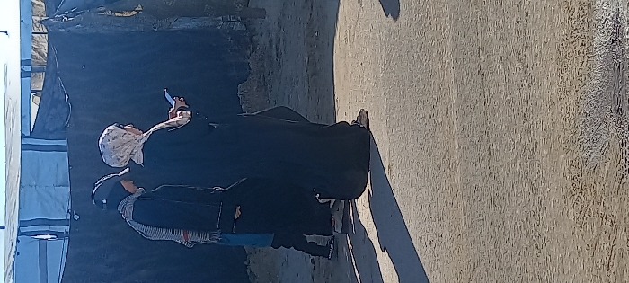 The image depicts two individuals walking on an uneven dirt or gravel surface under direct sunlight in Khan Yunis, Palestinian Territory. Distinct shadows are cast on the ground. In the background, makeshift shelters constructed from dark tarpaulin-like material and a lighter blue-and-white striped tarp are visible, indicating a temporary settlement environment.
The individual on the right is a female, seen from the side, wearing a full-length black abaya and a light-colored, subtly patterned hijab covering her head. She holds a white smartphone horizontally with both hands, appearing to view its screen. Her footwear consists of light-colored flat shoes or sandals.
The individual on the left is partially visible from behind, wearing a dark beanie, a horizontally striped scarf in light and dark tones around their neck, a dark long-sleeved garment or coat, and blue trousers. Their footwear consists of dark sneakers with white soles. Both individuals are positioned as if walking from left to right across the frame.