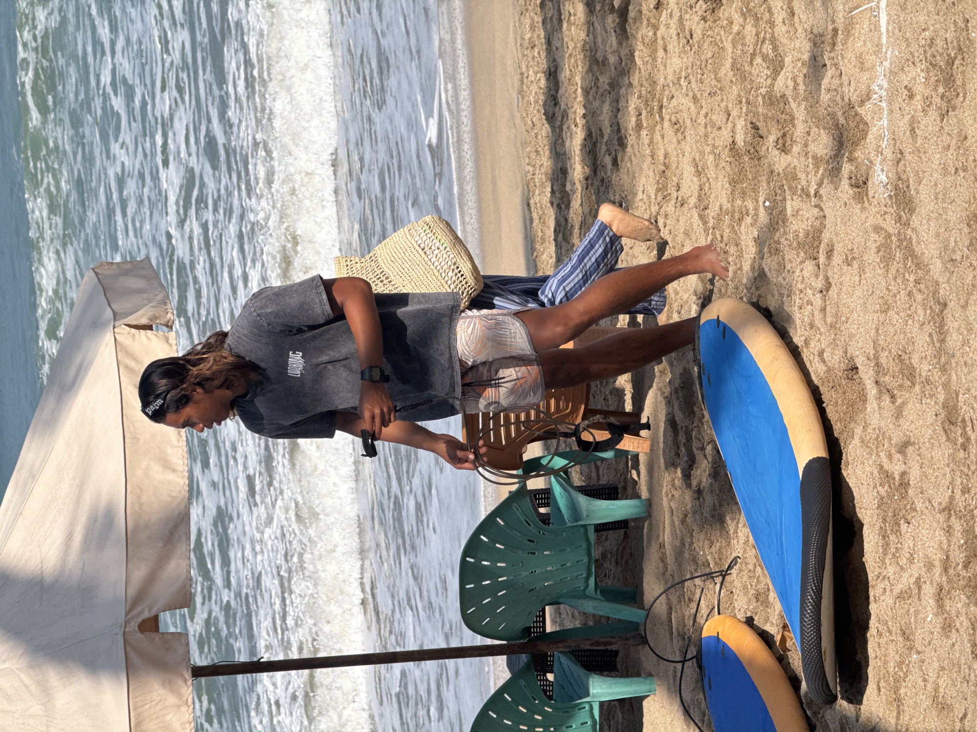 Two surfboards, one blue with orange trim and another predominantly blue with orange trim, lie on the sand at Kuta Beach, Indonesia. In the background, a person with long brown hair tied back with a black bandana is wearing a dark t-shirt and patterned shorts. They are holding a surf leash and a black object, possibly a GoPro. The person is standing next to a straw tote bag and is partially obscured by a beige umbrella. Behind them, two green plastic chairs are visible on the sand, and the ocean with moderate waves is in the distance. Another person, wearing blue and white striped pants and a tan sandal, is walking past. The sand is a light brown color.