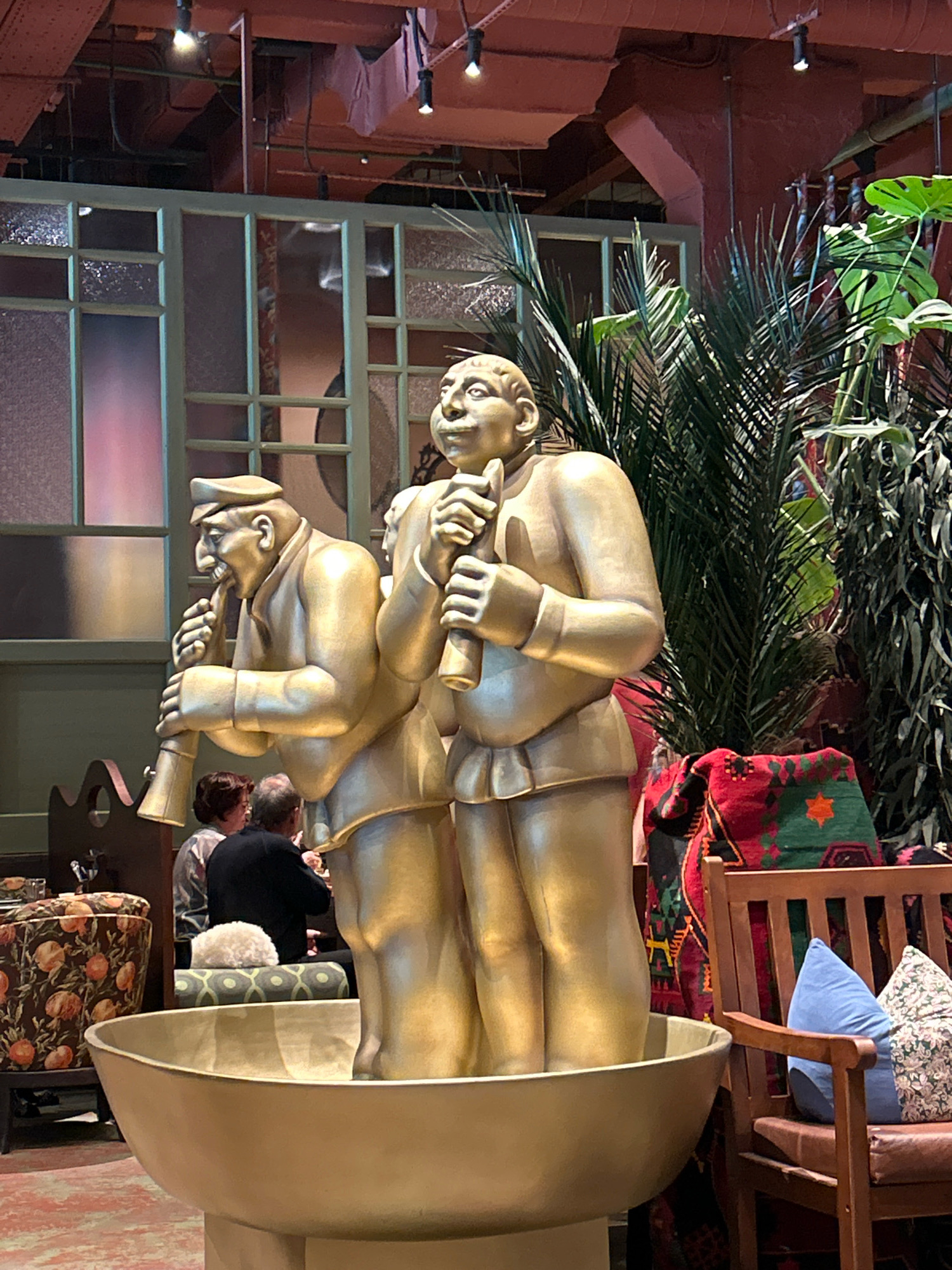 The image depicts a scene inside a restaurant with a dimly lit, bohemian ambiance. In the foreground, a large, golden sculpture of two men playing wind instruments dominates the frame. The sculpture is seated in a large, shallow golden bowl. Behind the sculpture, lush green plants, including palm fronds and large-leafed tropical plants, add to the exotic atmosphere. A wooden bench with colorful, patterned pillows and a red, green, and black woven blanket is visible on the right. In the mid-ground, patrons are seated at tables, with a blurred view of the restaurant's interior, including decorative chairs and stained-glass-like partitions. The overall impression is one of a unique and artistic dining establishment. The location is identified as Tomsk, Russia, though no specific restaurant name or further contextual information is provided within the image itself.