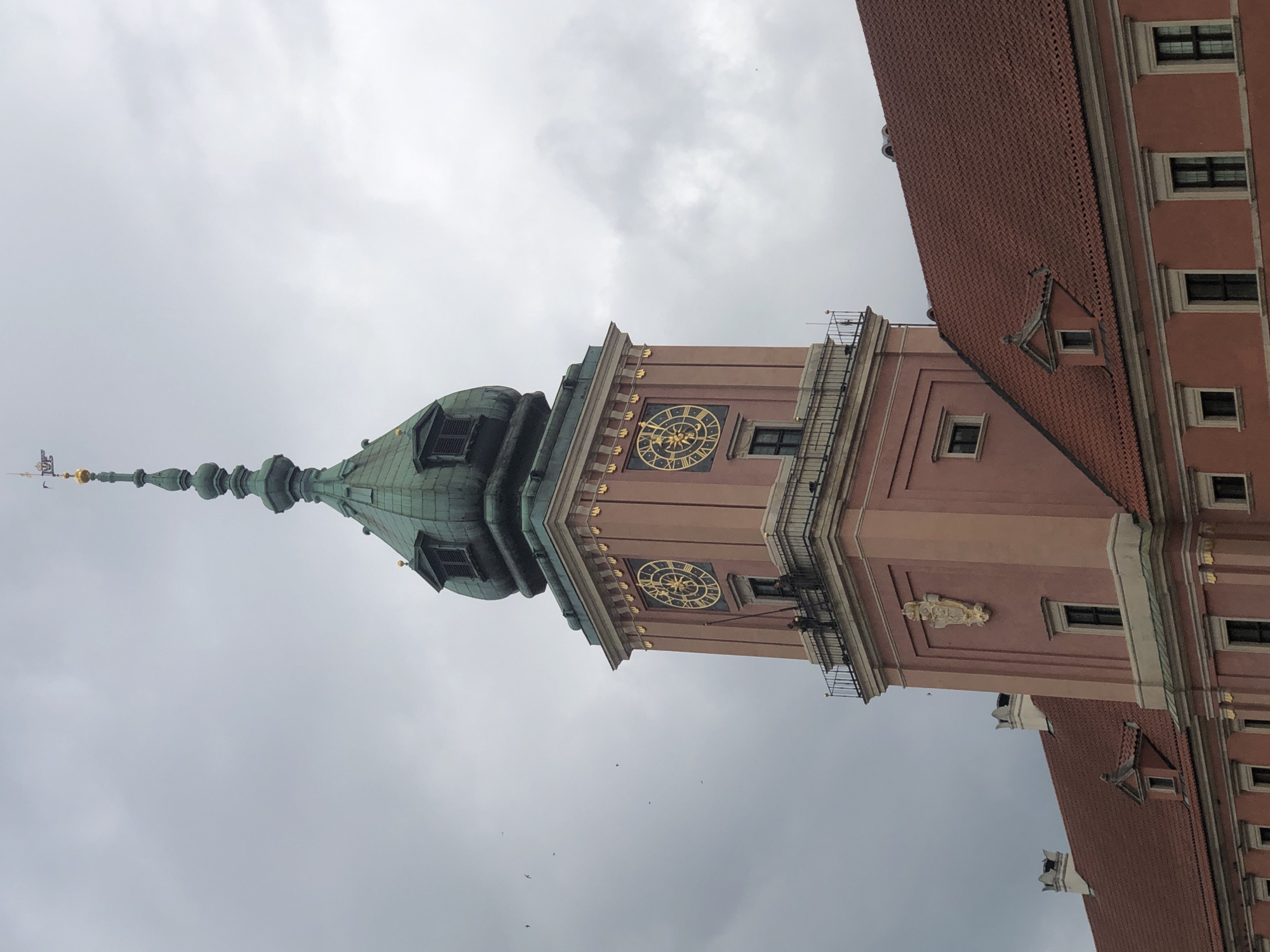 The image shows the Sigismund Tower (Zamek Królewski w Warszawie) located in Warsaw, Poland. The tower, painted in a muted pink hue, has a green, spired roof. Two ornate golden clocks are visible on the tower's facade, each with Roman numerals. A balcony with a railing is present below the clocks, and two figures, likely workers, are standing on it, one holding a long pole. The background consists of a cloudy, overcast sky with a few birds flying. The lower part of the image displays the tiled, red roof of an adjoining building, with visible windows and architectural details.