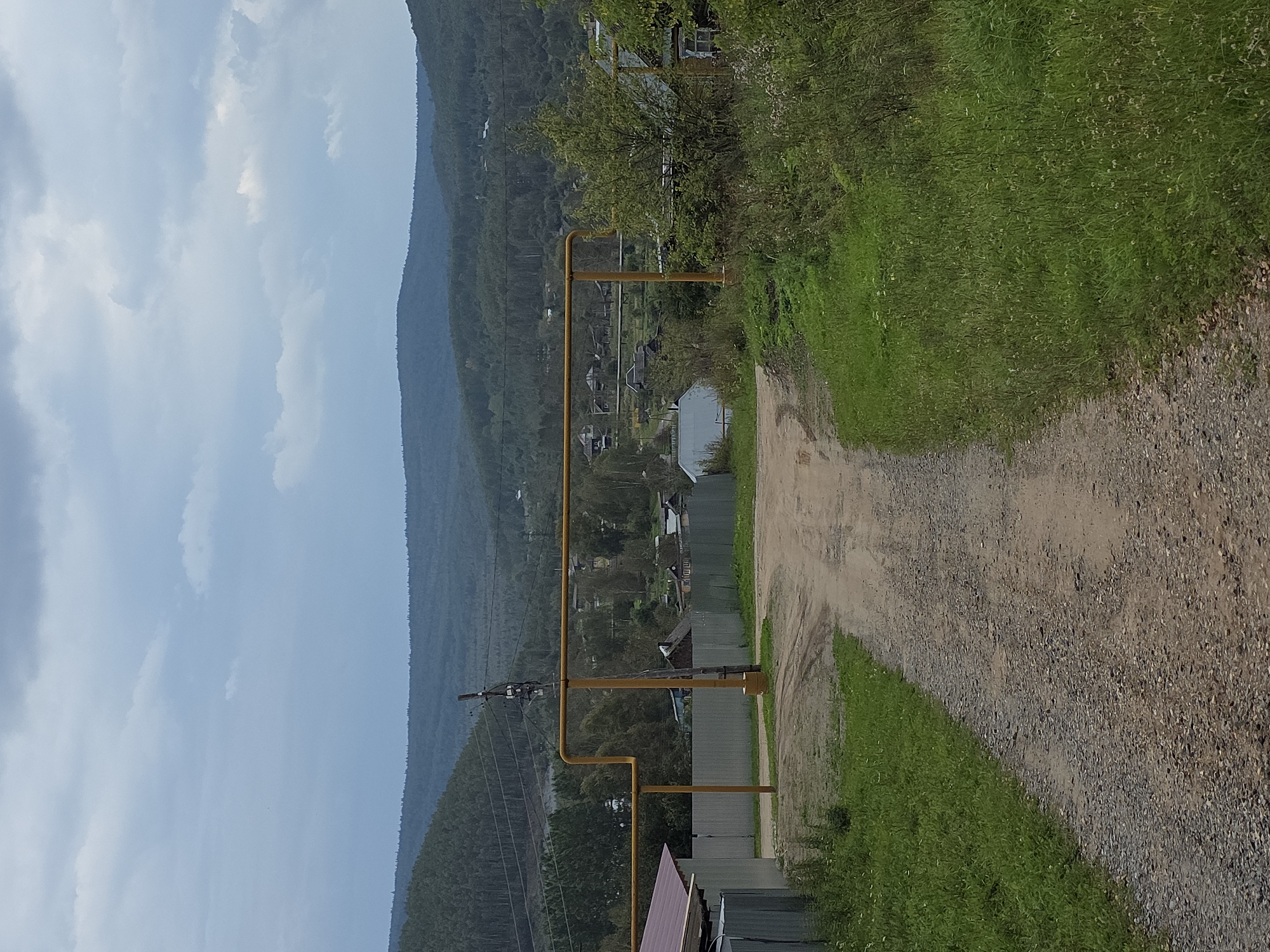 The image depicts a rural landscape near Magnitka, Russia, on an overcast day. A dirt road, worn by tire tracks, leads into the frame from the foreground and forks into two paths, one veering left and the other right. The left path is bordered by a corrugated metal fence and a building with a red-brown metal roof. To the right of the main road, a yellow gas pipeline runs horizontally across the scene, supported by vertical posts and a utility pole with electrical equipment. Beyond the pipeline, a village is nestled amidst trees and rolling hills. Houses with pitched roofs are visible, some appearing weathered. The background is dominated by densely forested hills that recede into the distance under a grey, cloudy sky. The vegetation consists of green grass and wild plants along the roadside and scattered trees. No people are present in the image.