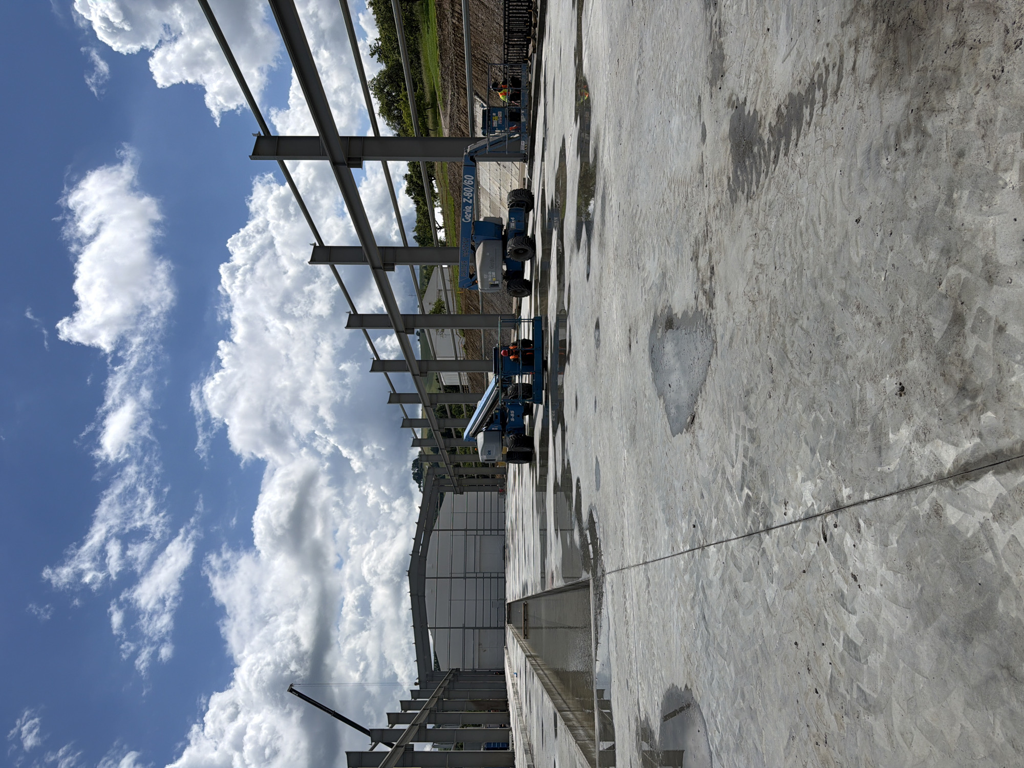 An outdoor construction site in Janowice, Poland is depicted. The scene shows a partially completed building structure with exposed steel beams. In the foreground, a wet concrete surface is visible with several puddles. A blue articulated boom lift, identified as a "Genie Z-60/60," is positioned in the middle ground. Two workers wearing orange vests are operating from the platforms of two different boom lifts. One boom lift is to the left and the other is to the right of the central boom lift. A large, unfinished building with a white facade stands in the background. The sky is bright blue with scattered white clouds. A construction crane arm extends from the left side of the image into the upper sky. The surrounding landscape includes a vegetated hill to the right.