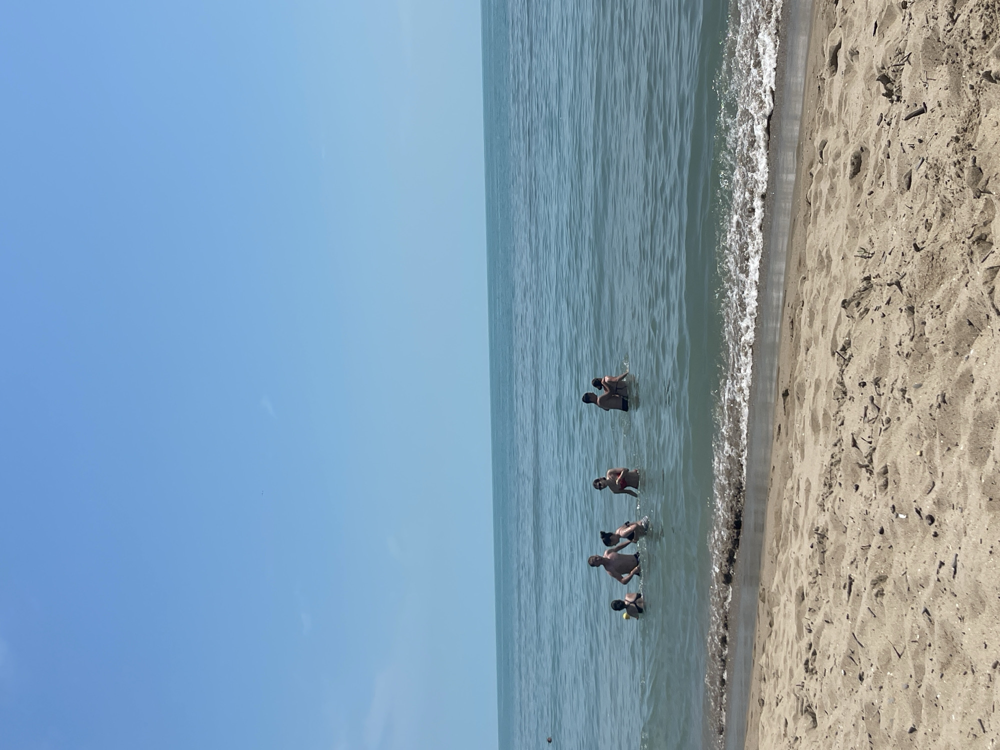 This image depicts a sunny beach scene in Sveti Vlas, Bulgaria, on a clear day. The frame is dominated by a bright, cloudless blue sky on the left, and the sea meeting a sandy beach on the right (when viewed in correct horizontal orientation).
Approximately six to seven people are visible in the calm, light blue-green waters of the sea, relatively close to the shore. They appear to be adults, mostly standing or wading with parts of their bodies submerged, suggesting they are enjoying a swim or cooling off in the water.
The sandy beach occupies the lower right portion of the frame, showing a textured, light brown surface with gentle waves breaking along the waterline. Some natural debris or small objects are scattered across the sand. The clear sky and bright illumination indicate it is daytime, likely morning or early afternoon, with pleasant weather conditions. The overall atmosphere is one of relaxation and leisure typical of a summer day at the seaside. There is no visible text in the image.
