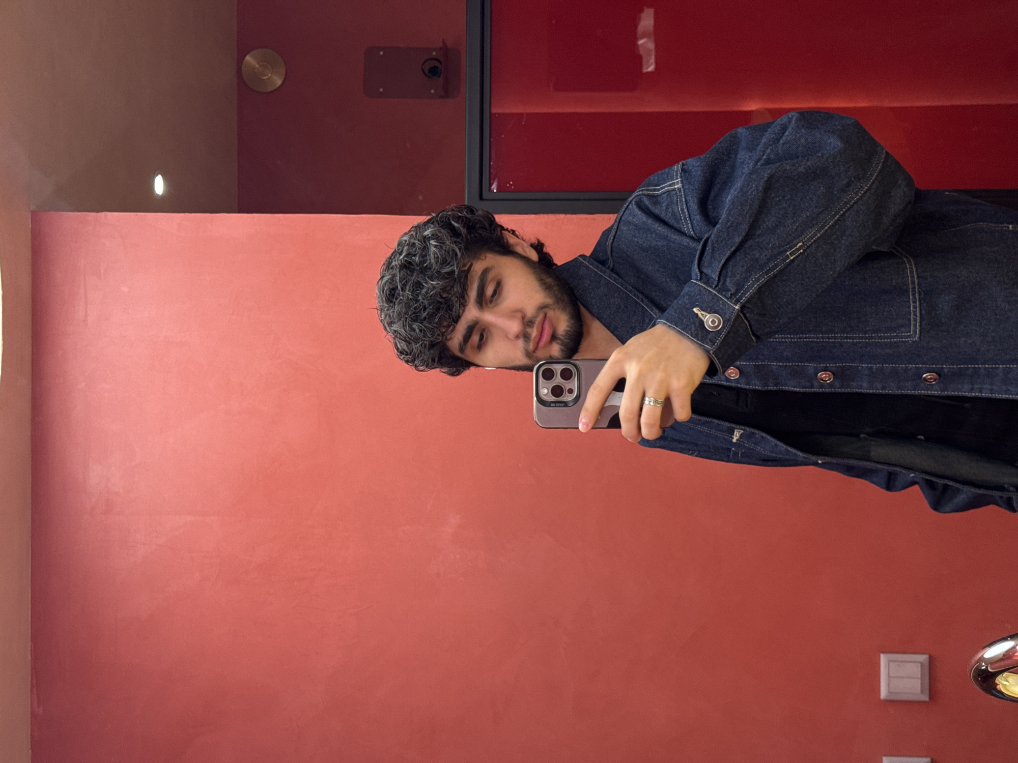 A young man with curly dark hair and a beard is taking a selfie in a mirror. He is wearing a dark denim jacket over a dark shirt. He's holding a grey smartphone with multiple camera lenses. He's wearing a silver ring. The background is a red-orange wall, possibly in a hotel or apartment. There is a light fixture and another red-painted section of the room above the wall. It is impossible to determine the precise location in Yerevan, Armenia, without further context.