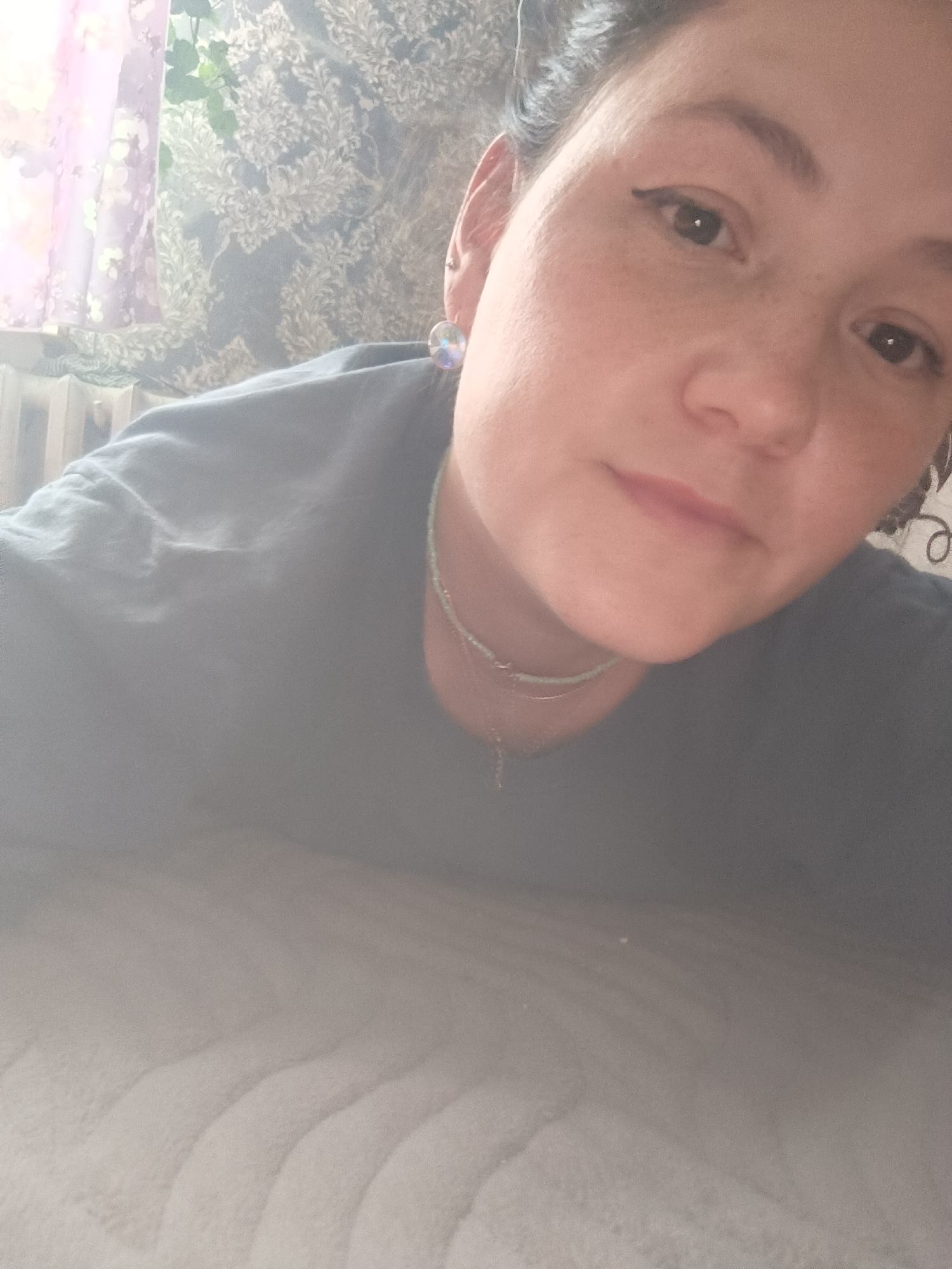 A close-up selfie of a woman lying down on a textured grey surface. She has dark hair pulled back and is wearing a dark grey t-shirt and a light turquoise beaded necklace with a small pendant. She has light makeup on, including winged eyeliner. Behind her, the background shows a window with sheer curtains in soft pink and white floral patterns, and what appears to be a radiator. The image is taken indoors in what could be a residential setting. The location is not specified as Karsha, Russia.
