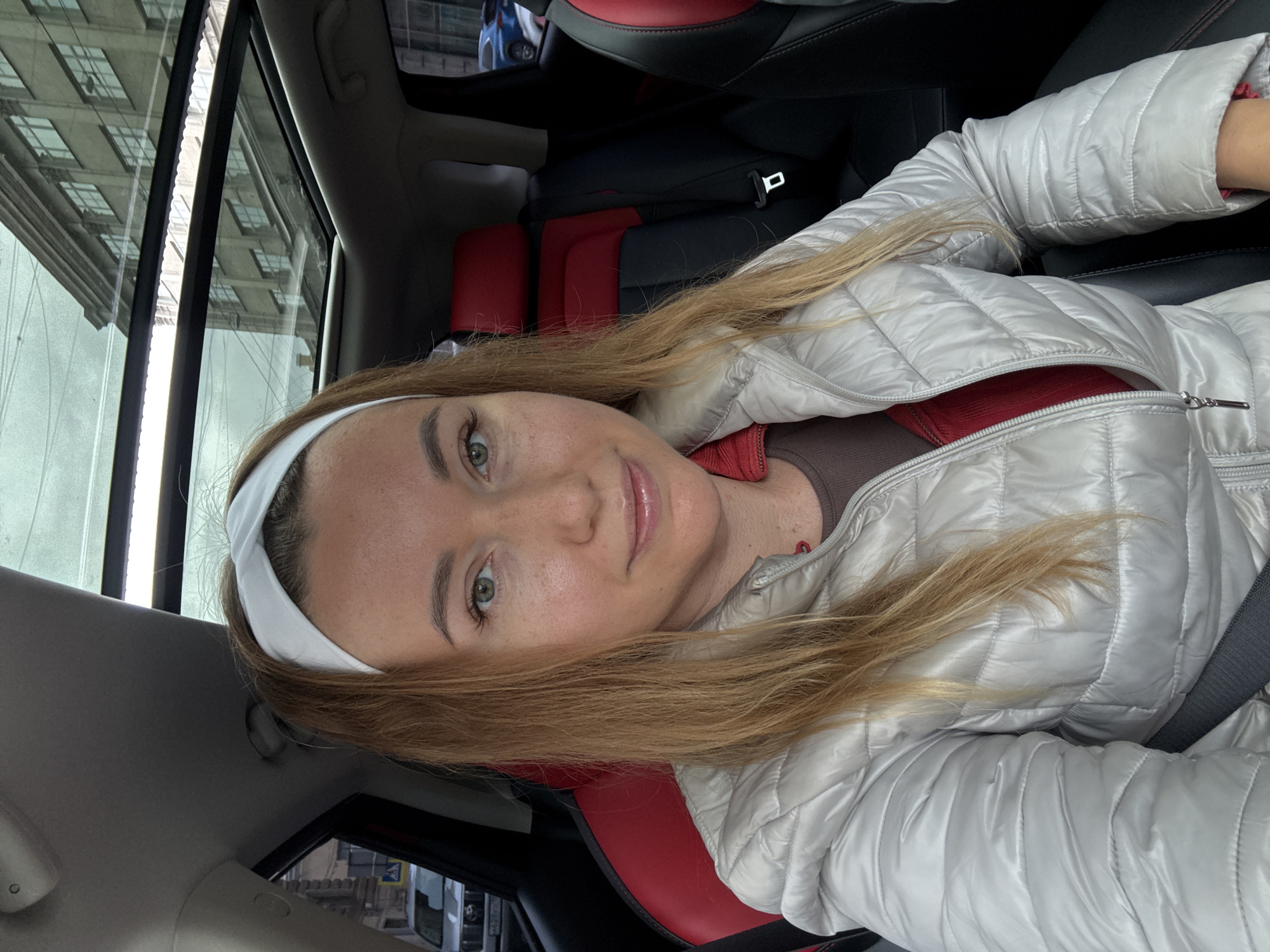 The image is a selfie of a Caucasian woman with long blonde hair and green eyes, wearing a white headband and a light grey puffy jacket over a red shirt. She is in a car, as indicated by the interior, including black and red seats and a visible seatbelt. Outside the car window, a cityscape is partially visible, implying an urban location. The city is not identifiable from the image.