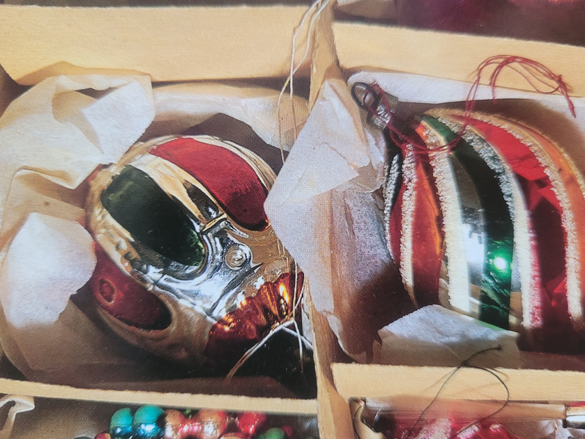 The image displays a close-up view of Christmas ornaments nestled in their packaging. The top ornament is a round, multi-faceted bulb with sections of red, green, and silver. It is partially wrapped in off-white tissue paper. Below it, another ornament, also round, features alternating stripes of red, white, and green, with a reflective surface. This ornament is also situated in protective packaging, with some red string visible. The background consists of cardboard dividers and the muted tones of the packaging material. The image provides no contextual information about the city of Gävle, Sweden, or any people or actions within the scene.