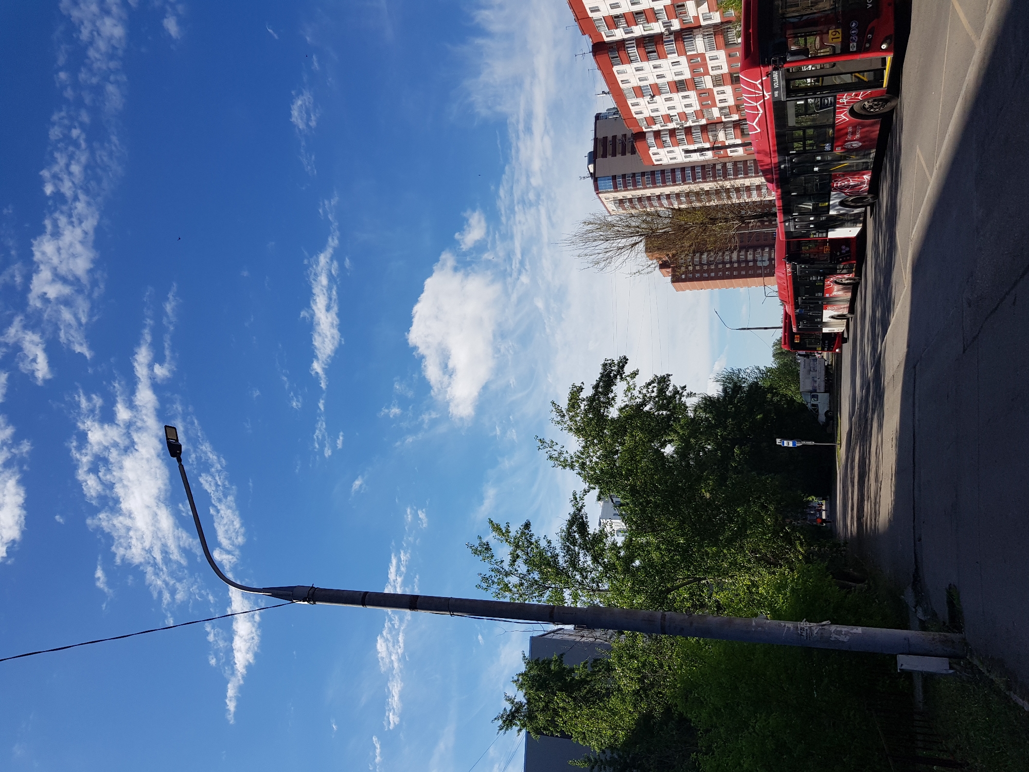 The image depicts a street scene in an urban environment. A row of red and white apartment buildings lines the street, with several red buses parked alongside the curb. A tree with green foliage partially obscures the view of the buildings. In the foreground, a tall gray lamppost extends upwards towards a blue sky with scattered white clouds. The street is paved and bordered by greenery on the side opposite the buildings.
