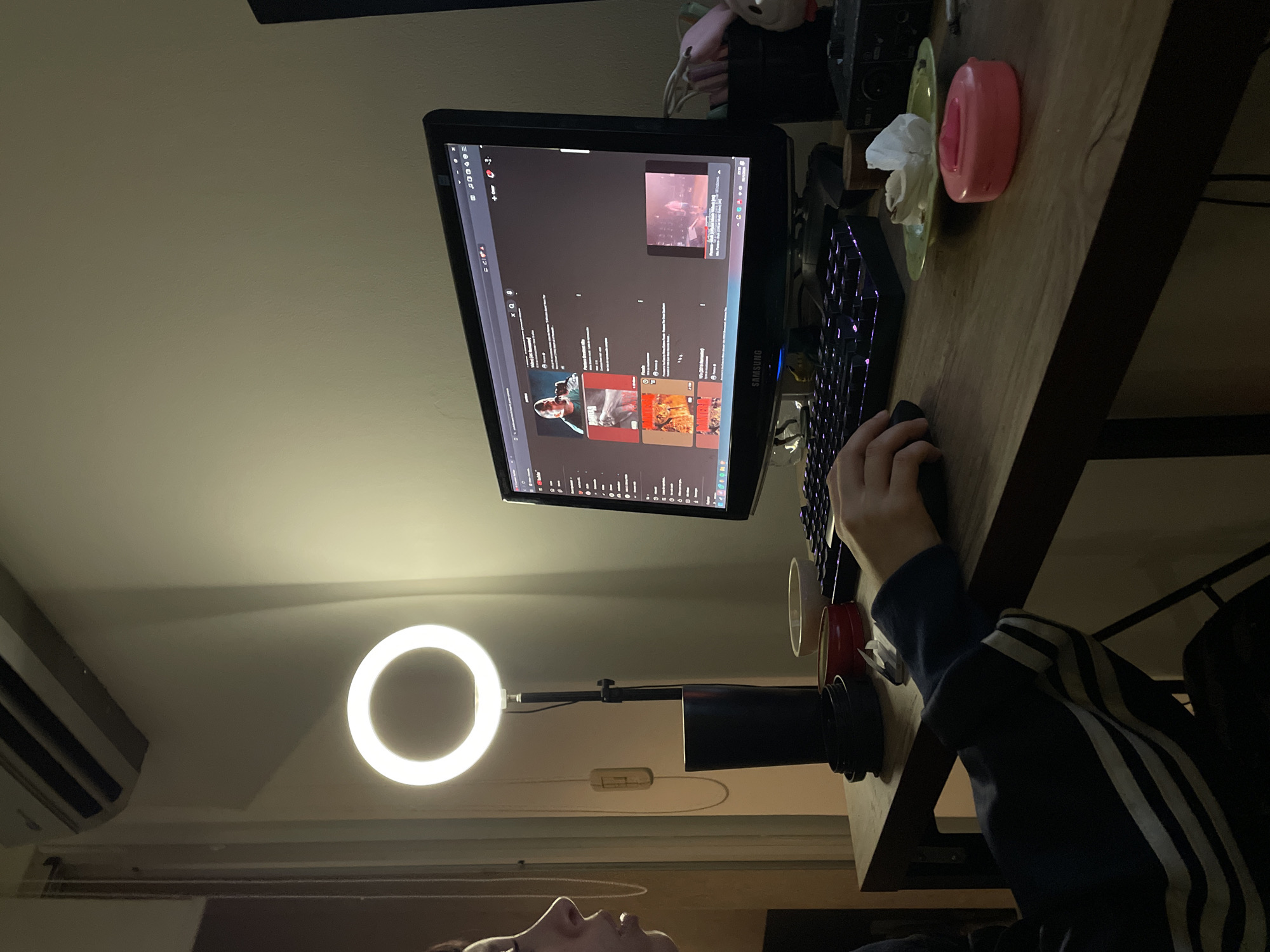 The image captures an indoor scene at night or in a dimly lit room, focusing on a person interacting with a computer setup. The person, whose lower face and right hand are visible, is seated at a wooden desk, wearing a dark blue sweatshirt with white stripes on the sleeve. Their right hand rests on a black computer mouse.
A Samsung monitor displays the YouTube interface, with a video playing in a smaller window at the top right, and various recommended videos below, including one titled "Phoenix". A black mechanical keyboard with purple backlighting is positioned in front of the monitor.
To the left of the monitor, a bright circular LED ring light stands on a tall, dark base, illuminating the immediate area and casting shadows. A red or dark-colored cup sits near the ring light. On the right side of the desk, next to the monitor, there's a pink object, possibly a tissue dispenser, with a white tissue protruding from it, resting on a small green plate or coaster. Additional wires and possibly audio equipment are partially visible towards the top right of the desk.
On the upper left wall, a white air conditioning unit is mounted. The overall lighting suggests an evening or nighttime setting, with artificial light sources dominating the scene. The person appears to be focused on the computer screen, possibly browsing content, streaming, or preparing for online interaction. The location context is Buenos Aires, Argentina.