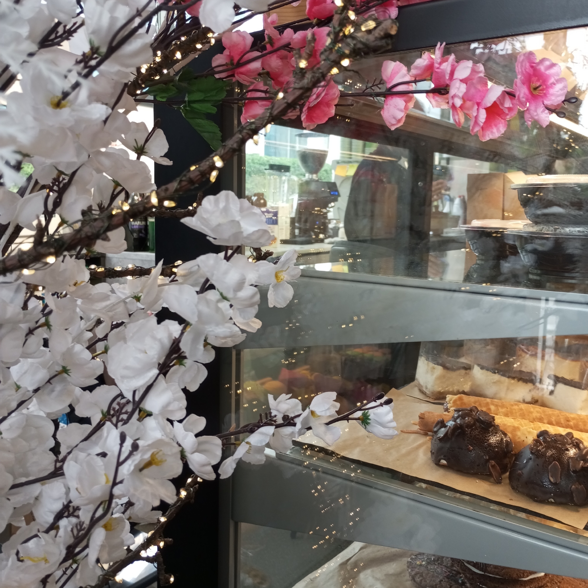 The scene depicts the interior of a commercial establishment, likely a cafe or bakery, located in Pervomaysky, Russia. In the immediate foreground, artificial flowering branches, predominantly white with some pink accents, are displayed, adorned with small, warm-toned LED string lights. These branches obscure a significant portion of the view.
Behind the decorative branches, a glass display case is visible, showcasing various food items. The upper section of the display case contains several dark-colored bowls, some with lids. The glass surface reflects portions of the establishment's interior and exterior; specifically, a coffee grinder or machine with blue indicator lights and a blurred reflection of a person, possibly an employee, are visible.
The lower shelf of the display case holds an assortment of desserts on a sheet of brown parchment paper. Prominently featured are two dark, chocolate-covered pastries, possibly truffles or small cakes, garnished with almond slivers. Behind these, several elongated, light-colored rolled pastries or waffles are arranged. Further back, larger, layered dessert slices or cakes are observable, featuring a light-colored base and a darker top layer. No direct human interaction is depicted, though the presence of the display case and its contents implies commercial activity.