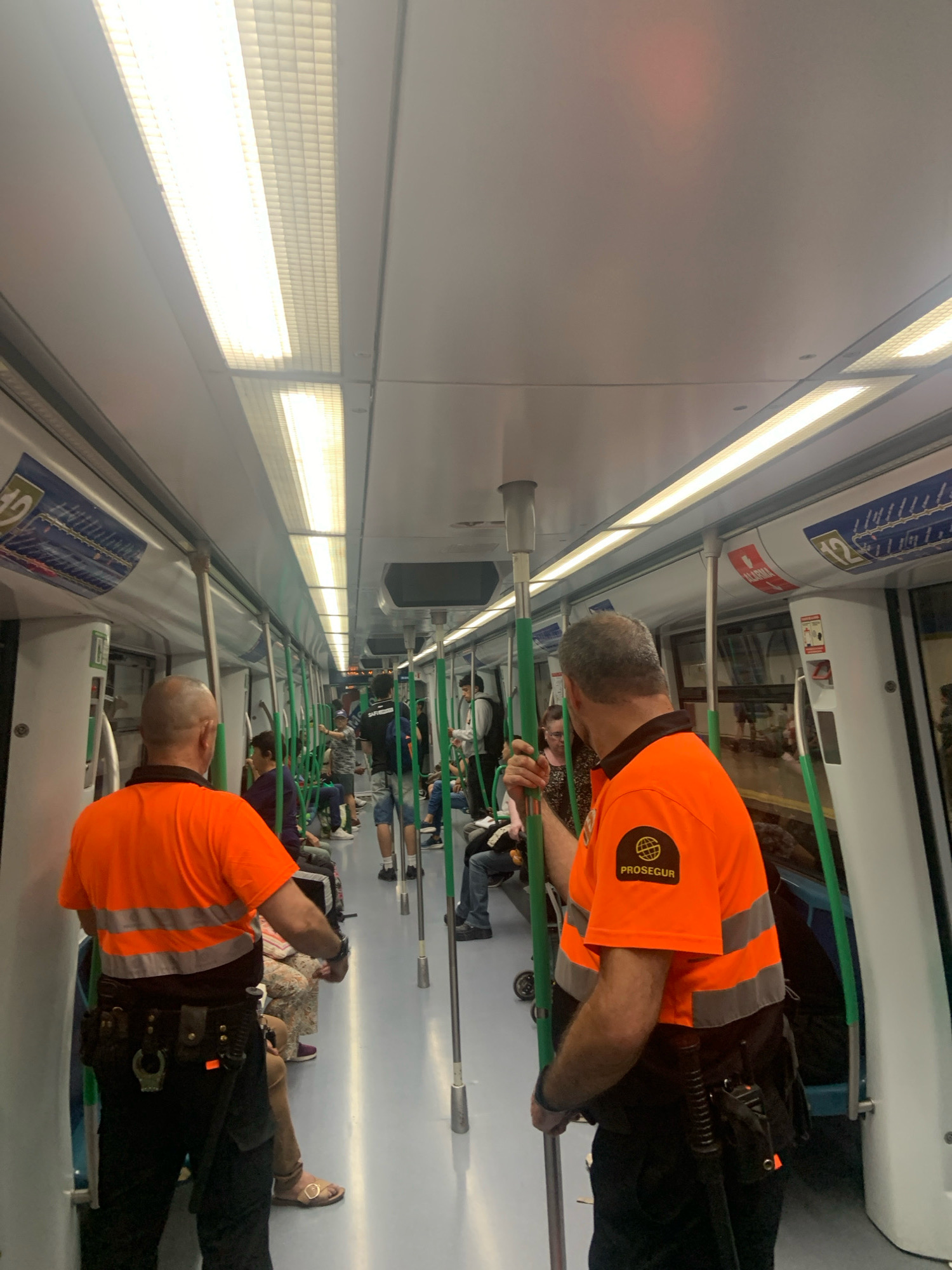 The image shows the interior of a metro train car in Alcorcón, Spain. Two men in orange safety vests with reflective stripes are visible in the foreground, facing away from the camera. The man on the left is wearing a black utility belt with a handcuff pouch and other equipment. The man on the right is also wearing a utility belt and has a patch on his vest that reads "PROSEGUR". They appear to be security personnel. Other passengers are seated and standing throughout the train car, some looking at their phones, others talking. The train car has white walls, fluorescent lighting, and green poles for passengers to hold onto. There are advertisements and signage on the walls. The overall scene depicts a typical commute on a public transport system.