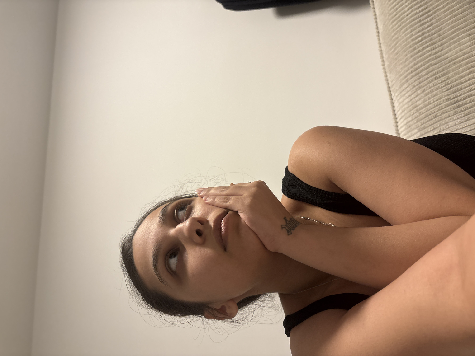 The image displays a young woman reclining indoors, captured from an overhead, tilted perspective. She is looking upwards and slightly to her left with a thoughtful or distant expression. Her right hand rests gently on her cheek, partially obscuring her mouth, while her arms are crossed in front of her. She wears a black top or dress with thin straps and a thin, silver-colored chain necklace. A tattoo is visible on her right forearm, appearing to read "Jadu" in a stylized script. Her dark hair is styled away from her face. The background consists of a plain, light-colored wall on the left side of the frame and a textured, light-colored cushion or upholstered surface on the right, suggesting she is on a bed or sofa. The lighting is soft and even, typical of indoor settings. No specific time of day or outdoor elements are discernible. The image provides no visual cues related to Mönchengladbach, Germany.