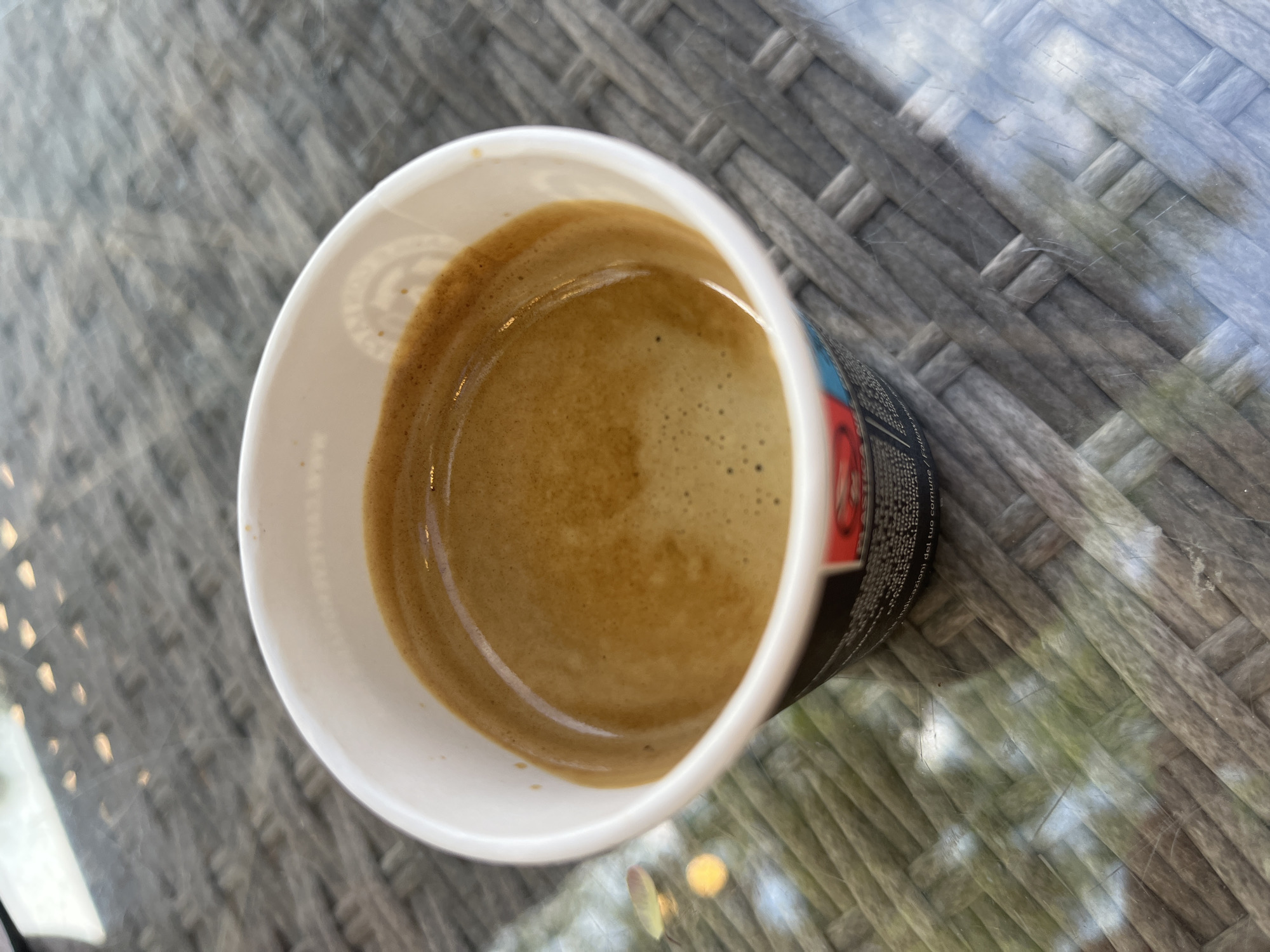 A close-up, high-angle shot shows a disposable coffee cup filled with dark, frothy espresso. The cup sits on a woven, grey surface, likely a table, with a textured, wicker-like pattern. Light reflects off the surface, suggesting it might be glass or a similarly reflective material. In the background, out of focus, hints of greenery and diffused light can be seen, possibly indicating an outdoor setting. The paper cup has some black text and a red graphic partially visible on its exterior. The location is Saint-Just-Saint-Rambert, France.