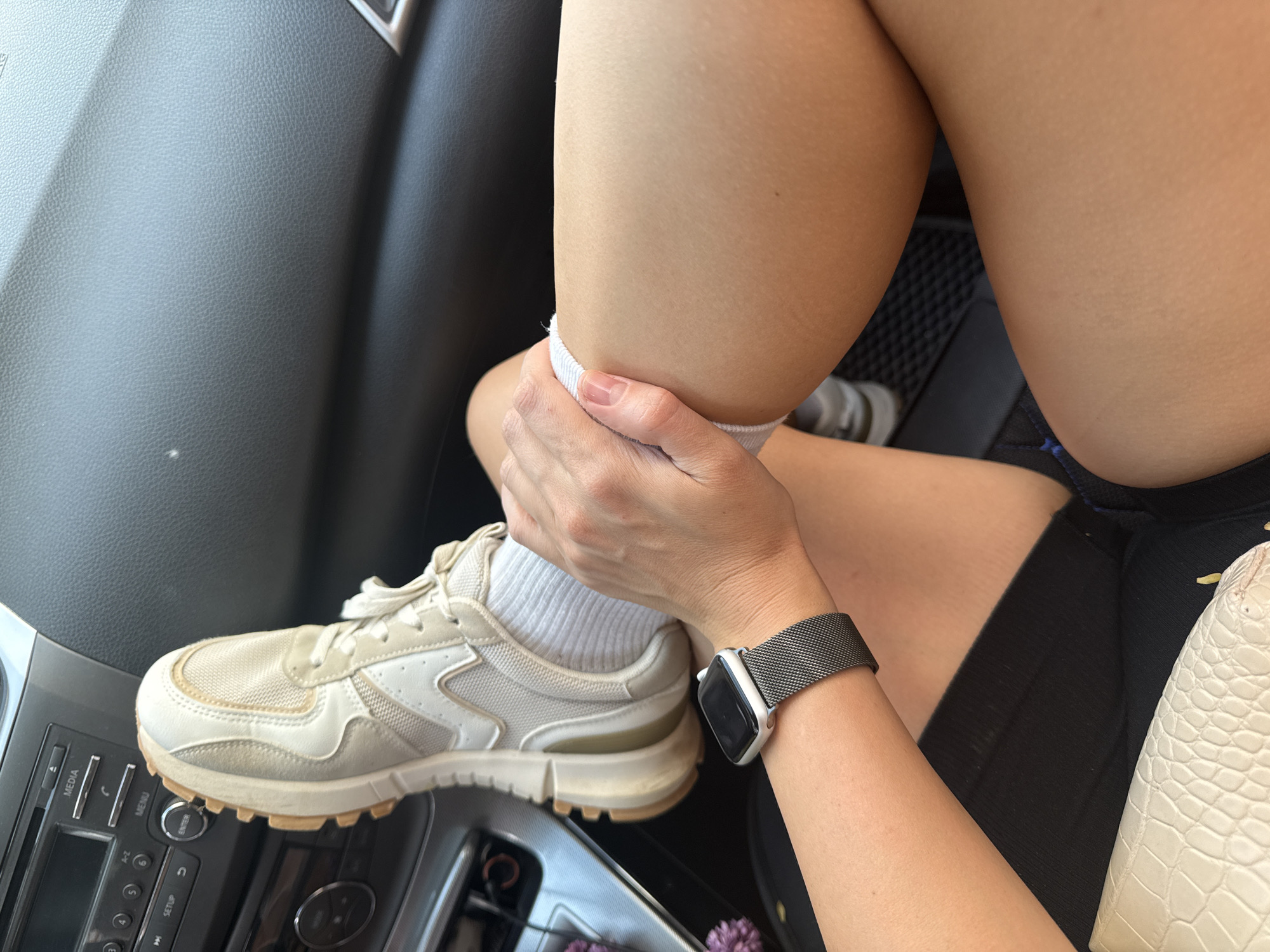 A person is seen sitting in the driver's seat of a car. The person's right leg is bent at the knee, with a white sneaker-clad foot resting on the car's dashboard. The person's hand is holding onto their calf just above the sock line. The person is wearing black shorts and an Apple Watch with a Milanese loop band. A white purse is partially visible next to the person's leg. The car's interior is visible, including the dashboard, climate control panel, and leather seats. The image does not provide enough information to determine whether this scene took place in the city of Chaikovsky, Russia.