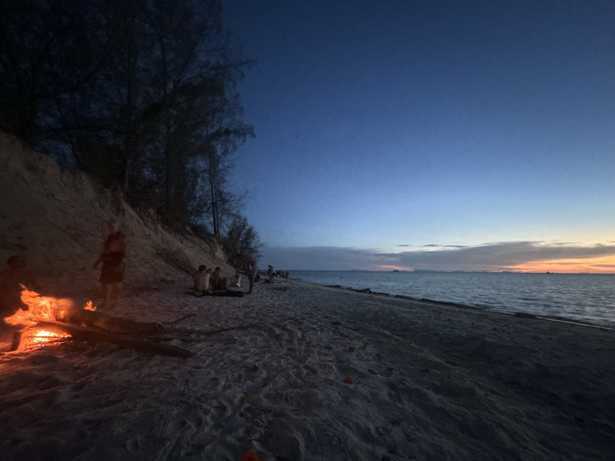 The image depicts a beach scene at dusk on Koh Phangan, Thailand. In the foreground, a bonfire burns brightly on the sandy shore. Several individuals are gathered around the fire, some appearing to be seated, while others stand. Behind the bonfire, a steep sandy embankment rises, with trees silhouetted against the darkening sky. The beach extends into the distance along the coast, meeting the calm sea. The sky is a deep blue, transitioning to hues of orange and yellow near the horizon, suggesting the sun has recently set. The overall atmosphere is one of relaxation and communal gathering at the end of the day.