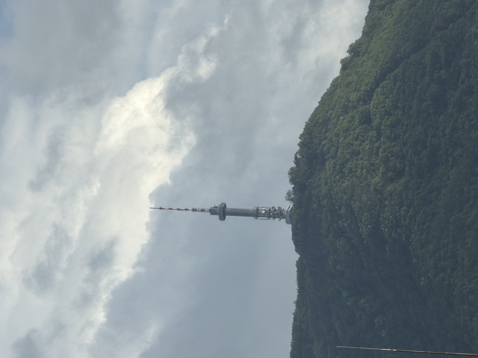 The image displays a tall communications tower positioned on a densely forested mountain under an overcast sky. The photograph is oriented vertically, showing the mountain slope rising steeply from the lower-right to the upper-right, while the sky and clouds dominate the left and upper-left portions of the frame.
The central subject is a robust, metallic communications tower, likely used for telecommunications or broadcasting. It features a broad, multi-tiered grey structure with various antennas and platforms, tapering into a slender mast that extends further skyward. This upper mast is marked with alternating red and white bands for visibility. The tower is anchored firmly into the terrain of the mountain.
The mountain itself is covered in a lush, dark green forest, indicating dense tree growth across its steep slopes. The natural contours of the land provide a dramatic backdrop for the man-made structure.
The sky is filled with a mixture of bright white and darker grey clouds, suggesting an overcast day. The lighting is soft and diffused, typical of a cloudy daytime setting, making it difficult to pinpoint the exact time of day. No specific activity or event is depicted, with the image focusing on the static presence of the tower and its natural surroundings. There is no visible text in the image. The scene captures a significant piece of infrastructure integrated into a rugged, mountainous landscape, characteristic of areas like Vitosha Mountain near Sofia, Bulgaria.