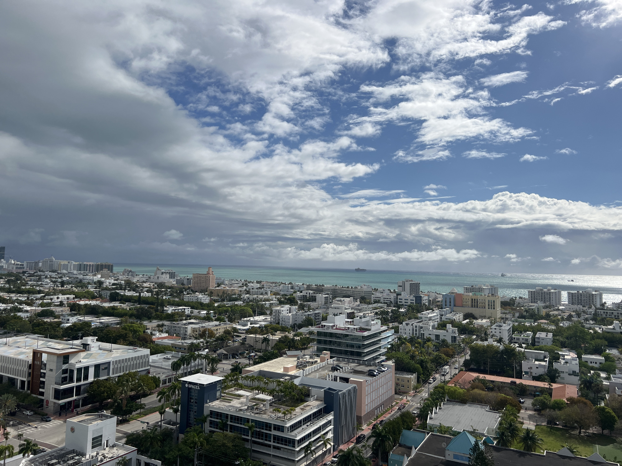 The image shows a cityscape in Miami Beach, Florida, USA, under a partly cloudy sky. In the foreground, a mix of residential and commercial buildings dominate the view, interspersed with green trees. The architectural styles vary, with modern, low-rise buildings with rooftop gardens and older, taller structures visible. The streets are lined with trees and some cars are visible. In the background, the Atlantic Ocean stretches to the horizon, reflecting sunlight. There is a ship or boat visible in the distance. The sky is filled with a mix of dark, heavy clouds and patches of blue sky with white clouds, suggesting an overcast day.