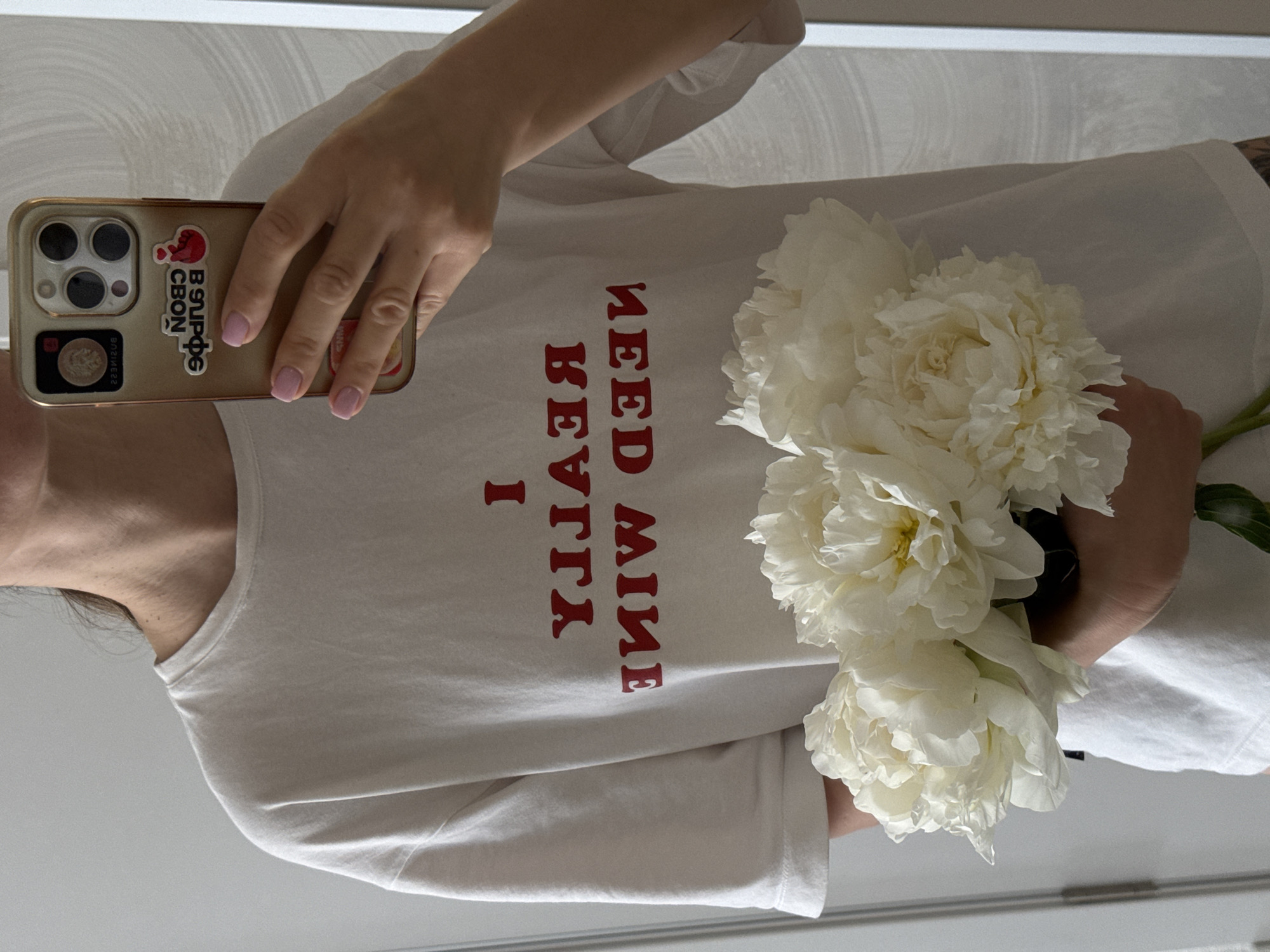 A person stands indoors, holding a bouquet of white flowers while taking a picture with a smartphone. The person is wearing a white T-shirt with the reversed text "I REALLY NEED MINE." The phone has a gold case with various stickers. The background appears to be a simple, neutral-colored wall. There is no indication of Sochi, Russia.