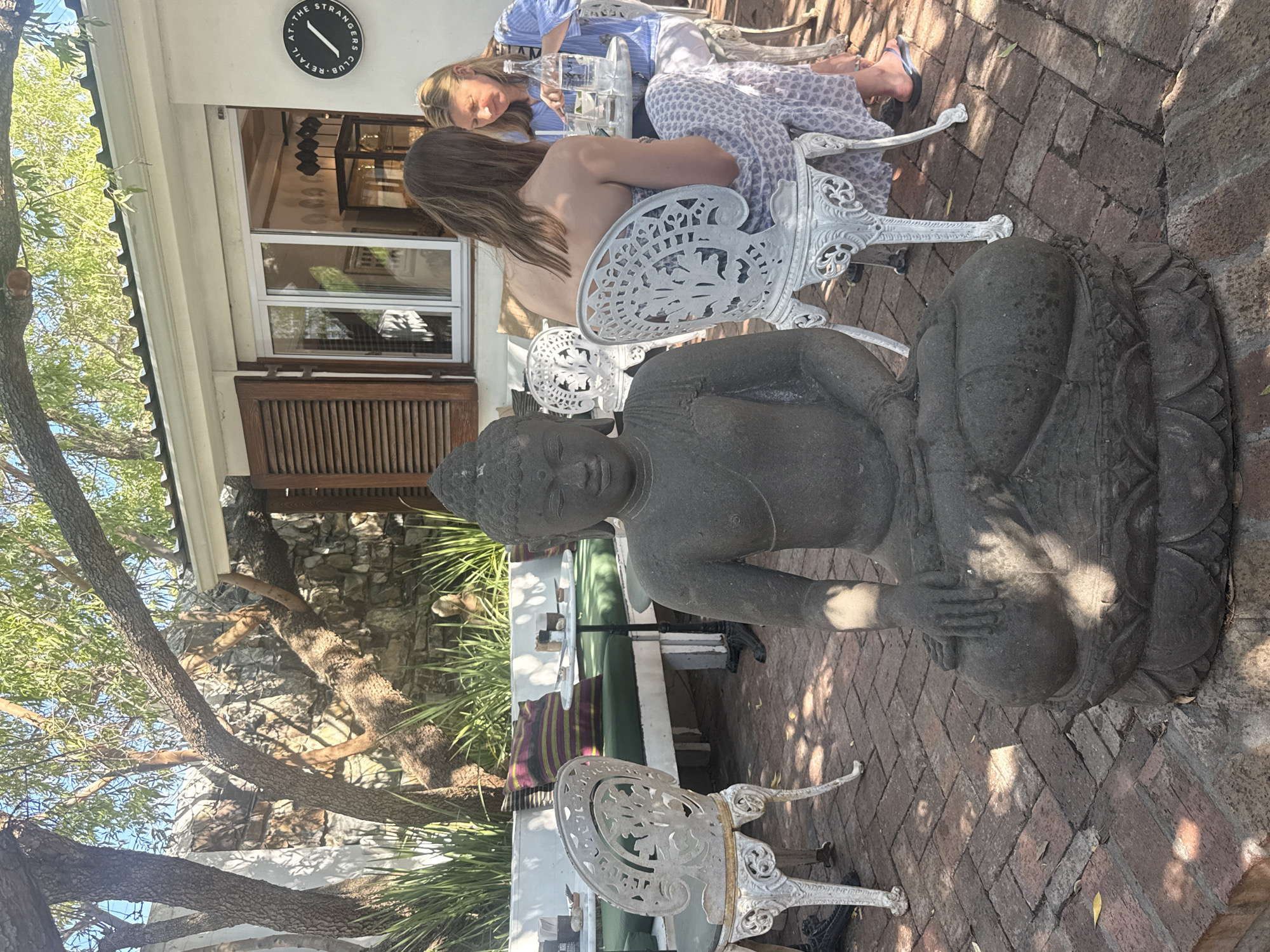 A photograph depicts an outdoor scene, likely a patio or garden area of a cafe or restaurant, possibly in Cape Town, South Africa, given the building style and foliage. The foreground is dominated by a stone Buddha statue, positioned centrally. Behind the statue, two women are seated at a table, appearing to be engaged in conversation and drinking from glasses. The furniture is painted white with ornate designs. The background includes a building with shuttered windows and a sign with "AT THE STRANGER'S CLUB" written on it. Brick paving makes up the ground of the patio area. Trees and foliage are visible on the periphery.