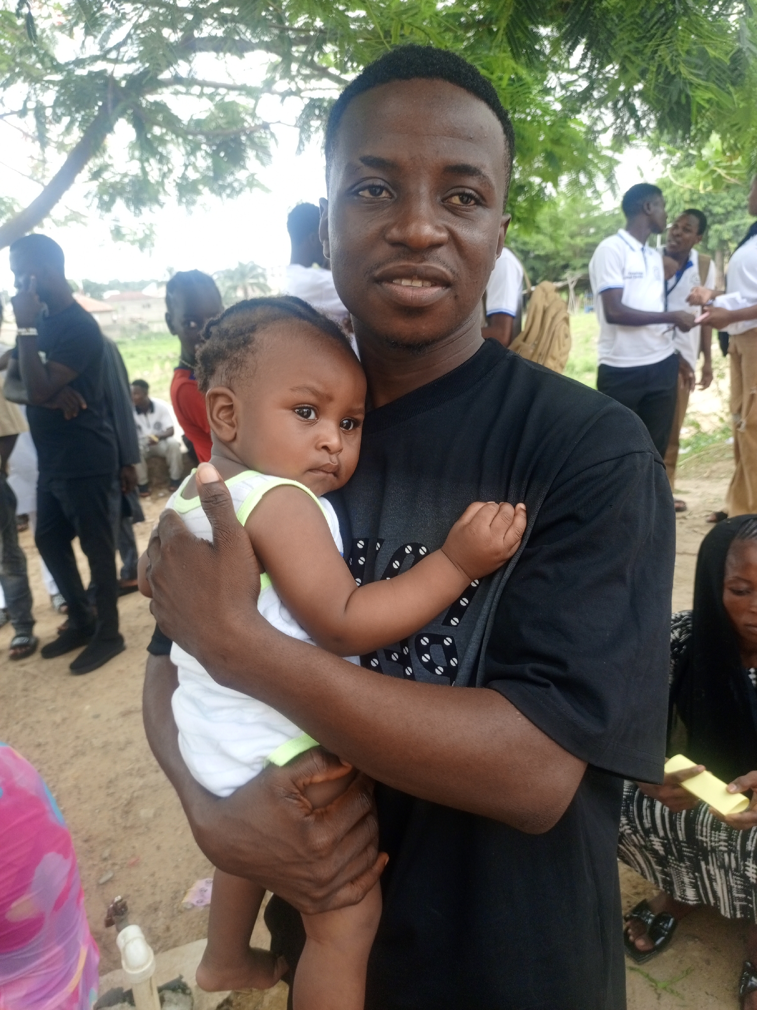 A medium shot of a dark-skinned man holding a baby, possibly a young child, outdoors in an unidentified urban setting. The man is wearing a black t-shirt. The baby is dressed in a white sleeveless shirt with green trim. The background includes other people, some wearing white shirts and khaki pants, and trees. The overall environment suggests a public gathering or event. It is daytime with outdoor lighting. There are some buildings visible in the background. The location is Unknown, Unknown.