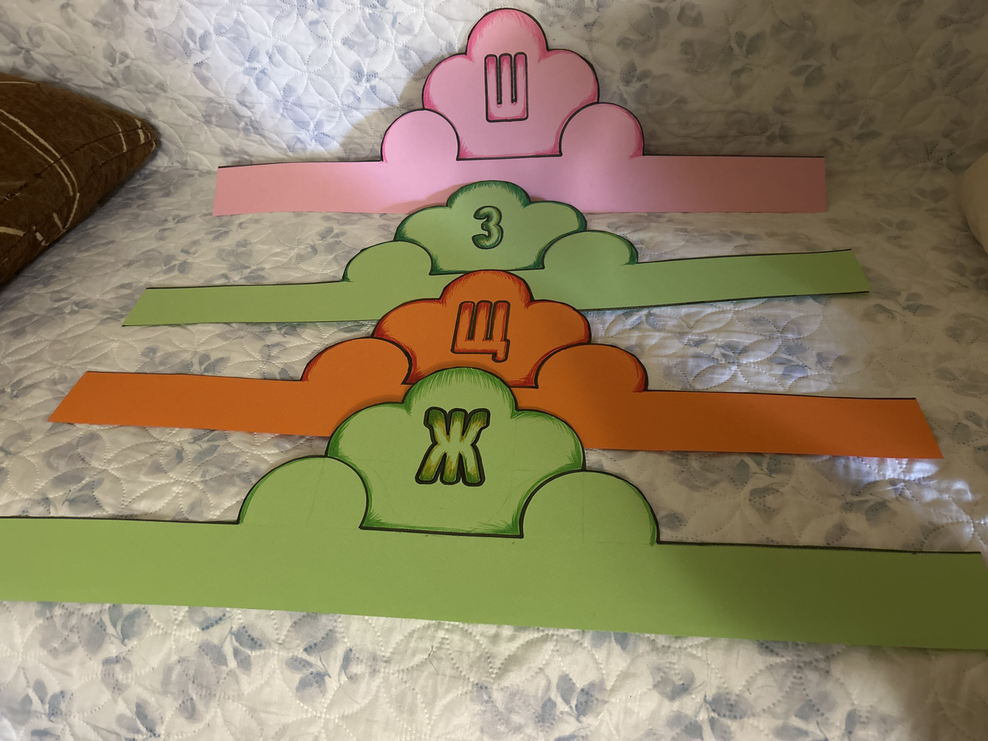 The image shows four paper crowns in a row on a patterned surface, possibly a bedspread. The crowns are cut in a cloud shape on top with a flat band for the head. From top to bottom, the crowns are pink with a Cyrillic letter "Ш" inside the cloud; light green with the number "3" inside; orange with a Cyrillic letter "Щ" inside; and darker green with a Cyrillic letter "Ж" inside. The crowns are outlined with black marker. There is a brown pillow visible on the left side of the frame. There are no indications of this image being in Cherven, Bulgaria.