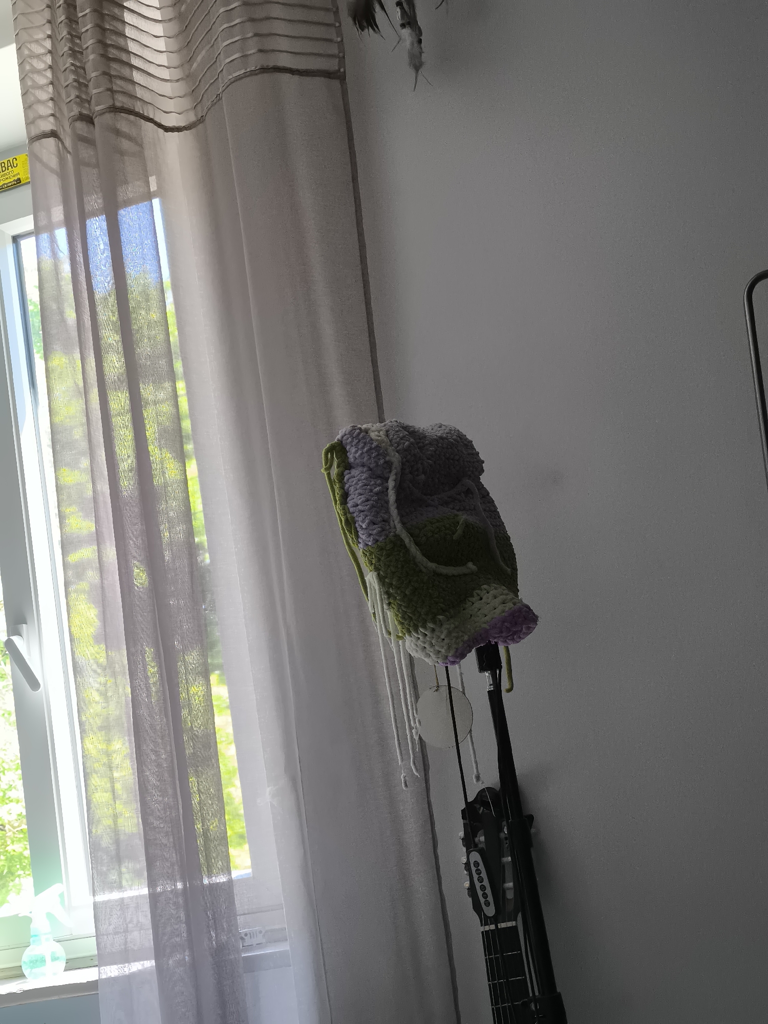The image shows an indoor scene with a window to the left covered by sheer, vertically striped curtains. Sunlight streams through the window, illuminating the interior. Outside the window, green foliage is visible.
To the right of the window, against a light grey or white wall, a guitar hangs, on which a colorful crocheted bag rests. The bag appears to be purple, green, and white. White cords or tassels hang from the bag.
The guitar is black and appears to be an acoustic model. The scene is lit by natural light, and the overall environment suggests a domestic setting.
There is no indication that the scene is located in Pélussin, France.