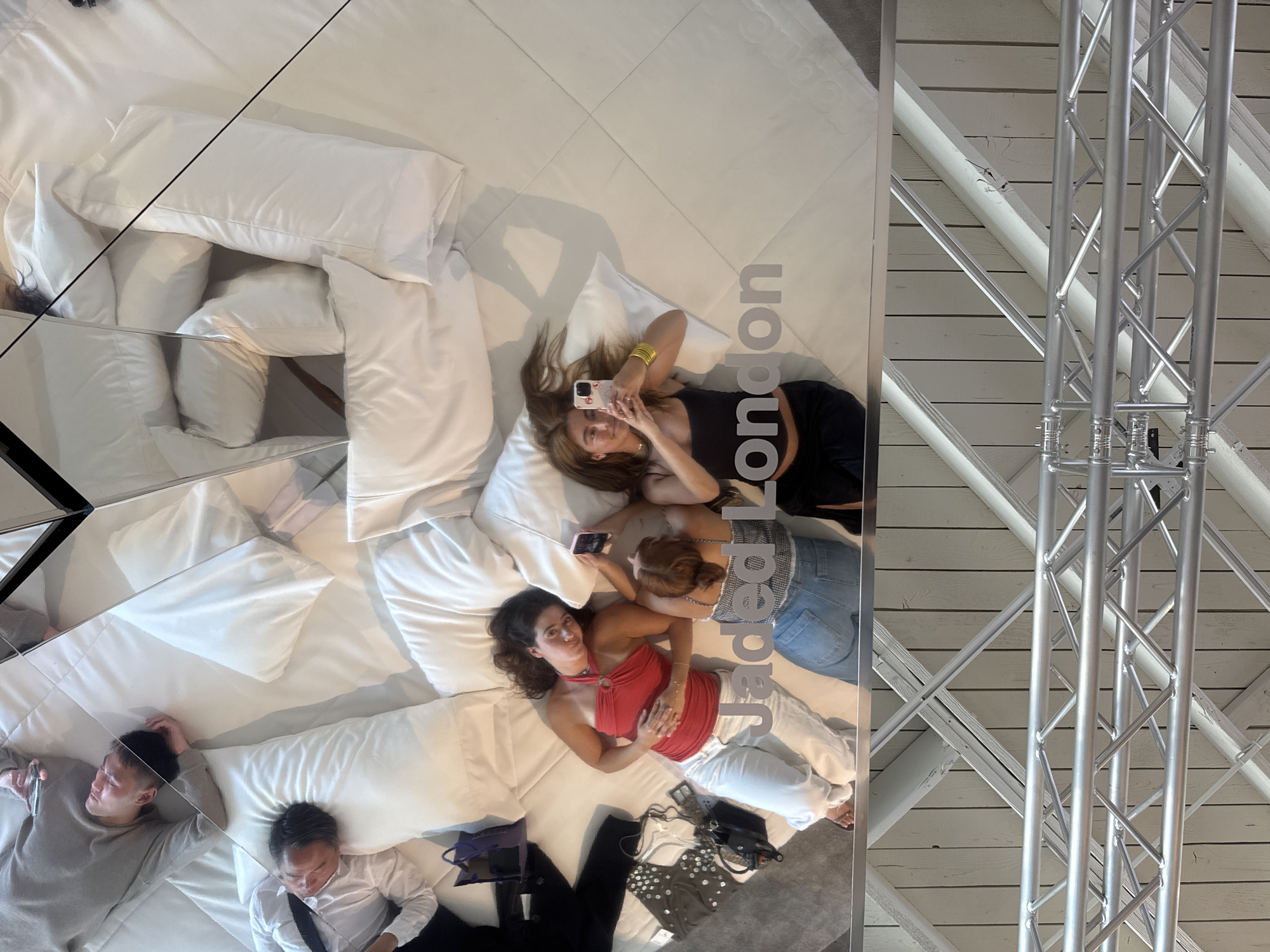 The image captures an overhead perspective within a structure in West Hollywood, USA, featuring a unique setup of interconnected mirrors that reflects a scene of several people casually lying on a mattress with white pillows. Two women are captured taking photos with their mobile phones. Adjacent to the mattress are personal belongings such as bags and clothes. The reflective nature of the mirrors distorts the view, creating multiple angles of the subjects and objects within the space. The interior of the building is constructed with light-colored wooden beams, crossed by silver metal trusses.