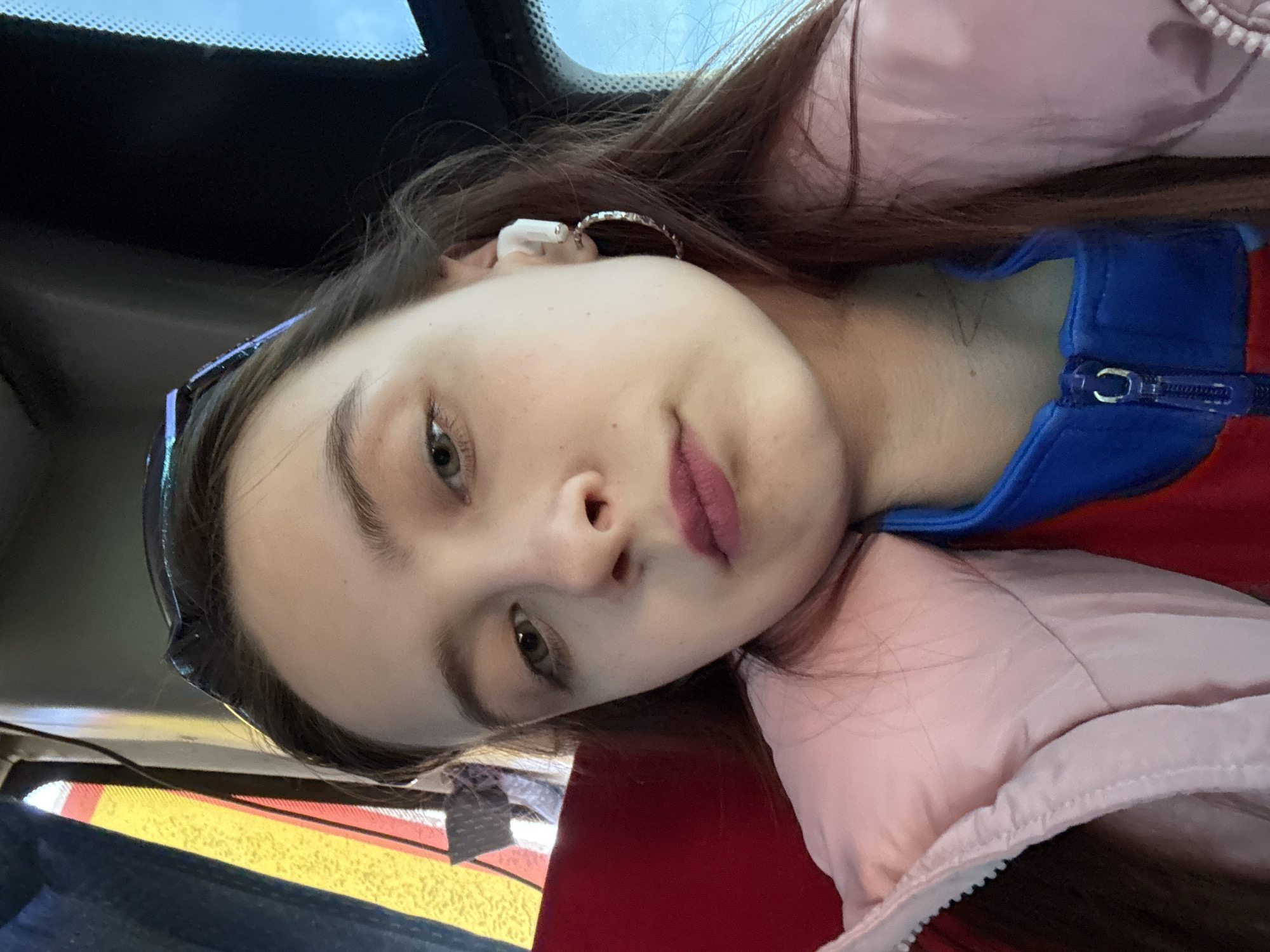 A young woman is in a car in an unknown location, but she is not located in the city of Usolye, Russia. She has fair skin, brown hair, and is wearing a pink jacket over a red and blue top. She is wearing an earring and a white earbud in her left ear. Sunglasses are perched on her head. The interior of the car is visible, including the dashboard and a glimpse of the windshield. The sky is partially visible through the windshield.