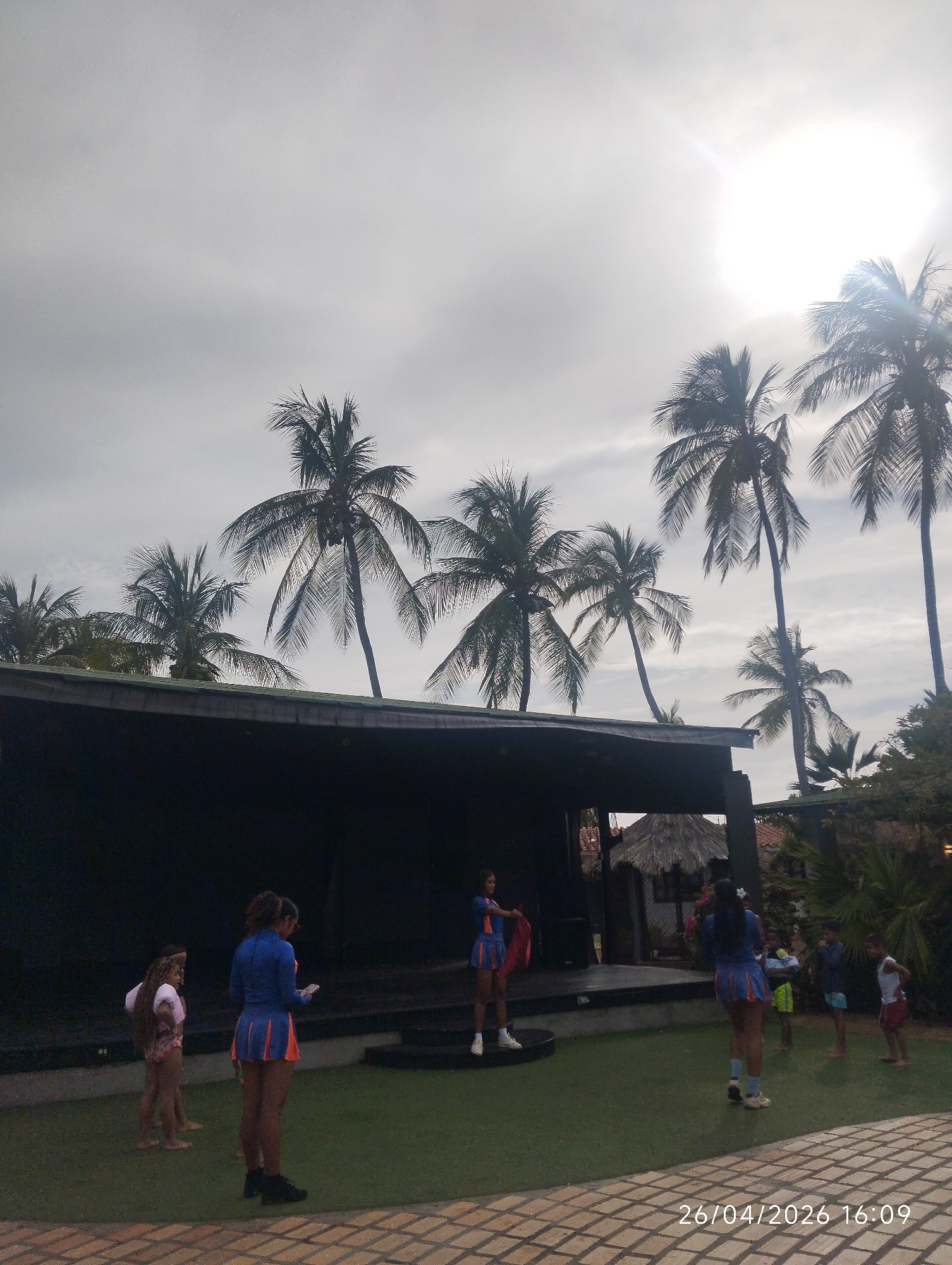 The image depicts an outdoor stage scene in what appears to be a tropical resort setting. Several tall palm trees stand behind the stage, silhouetted against a bright, overcast sky. In front of the stage, a group of people, mostly young women, are gathered on an artificial turf lawn. The young women are wearing matching blue and orange outfits. One person on the stage is holding a red piece of fabric. Other people are present, standing around the group. The surface in the foreground is composed of patterned pavement stones. The timestamp 26/04/2026 16:09 is visible in the bottom right corner. Given the city reference, the likely location is Porlamar, Venezuela. A thatched-roof structure is visible to the right of the stage, and some buildings are visible in the background.