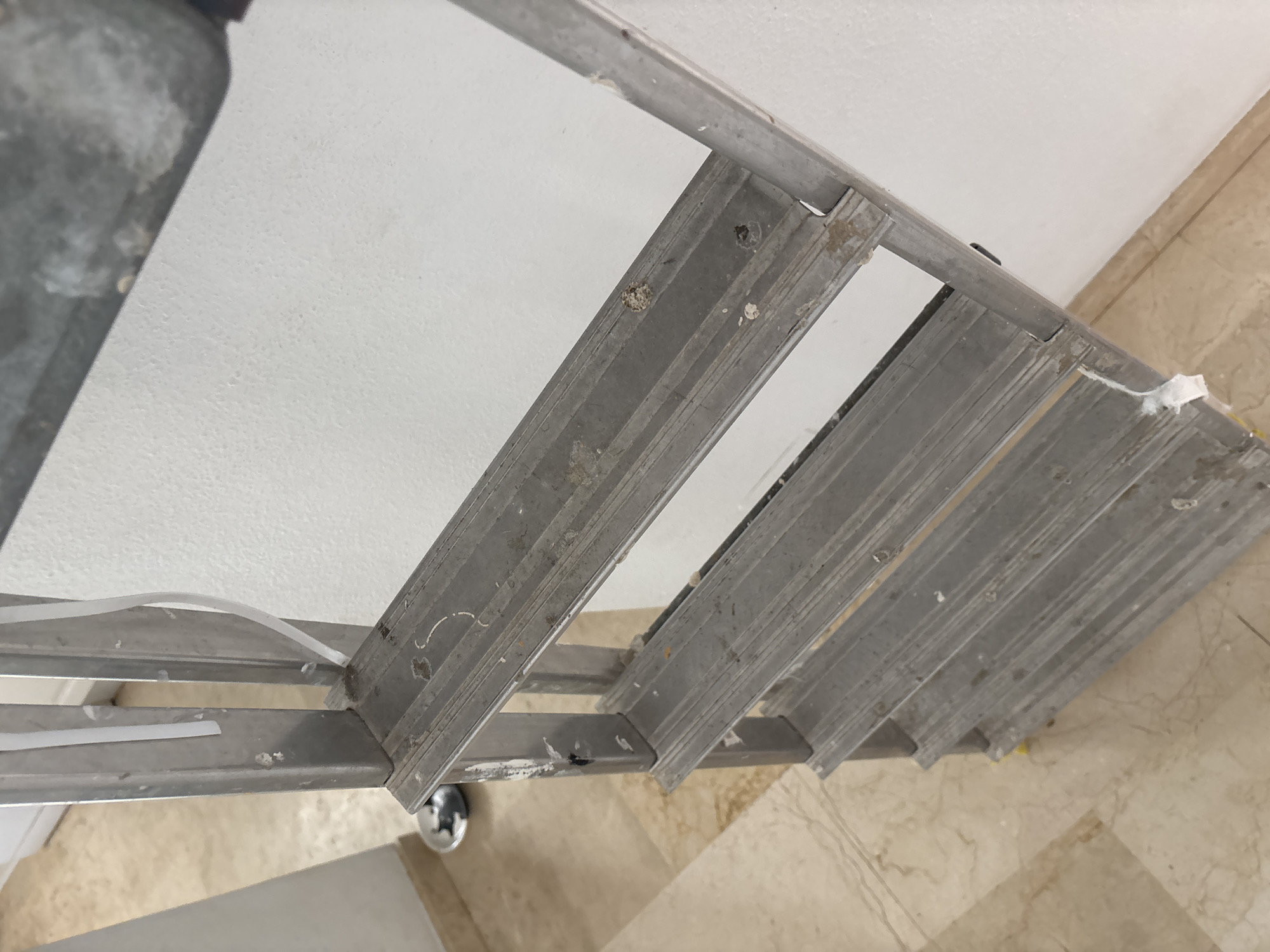 The image shows a close-up, low-angle view of a silver-colored metal stepladder leaning against a white textured wall. The ladder has at least four visible steps, all made of ribbed metal. There are some white debris, possibly paint or plaster, adhered to the wall and the ladder. A thin white cable or strip is attached to the ladder's side rails in multiple places. The floor is covered with light-colored marble tiles. The scene appears to be indoors, possibly in a room that is being renovated or maintained, and the overall impression is one of utilitarian use and recent activity. There are no people or animals visible in the image. The location is not specified within the image itself, and therefore its presence in Orihuela, Spain cannot be determined from the visual information alone.